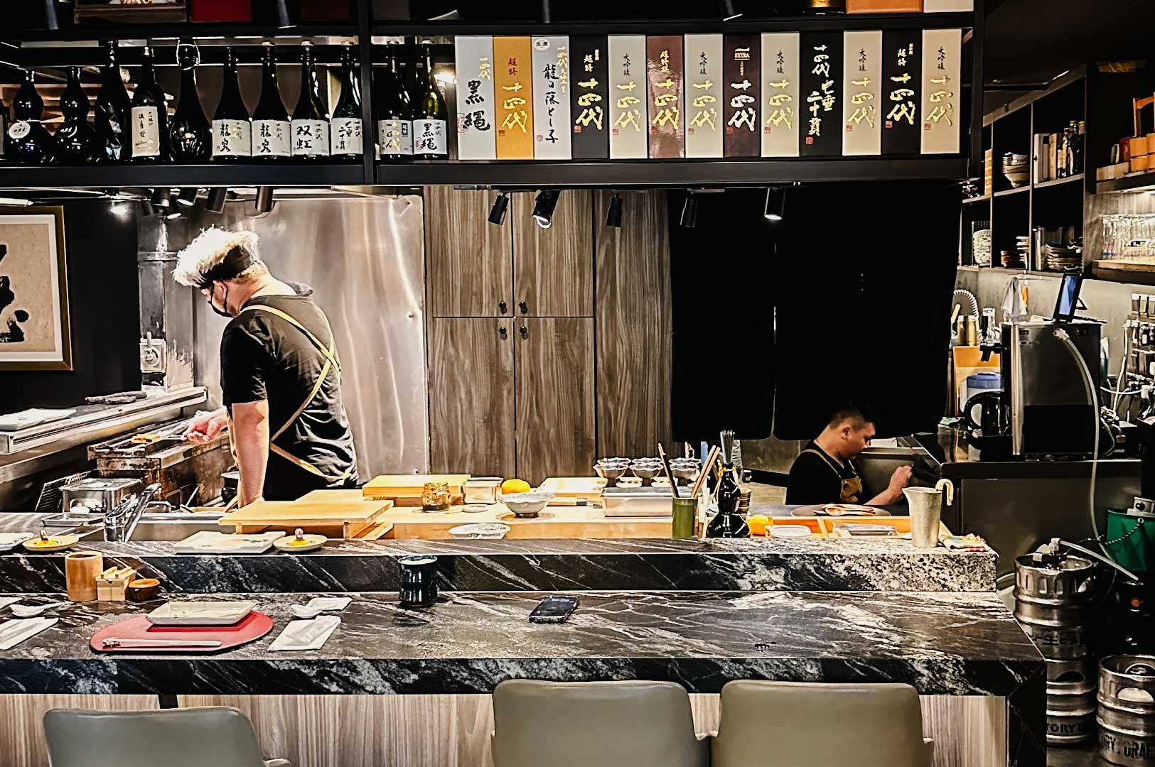 Japanese omakase-style counter with marble finishing, shelves of sake bottles overhead, and chefs preparing dishes behind the neatly arranged workstation.