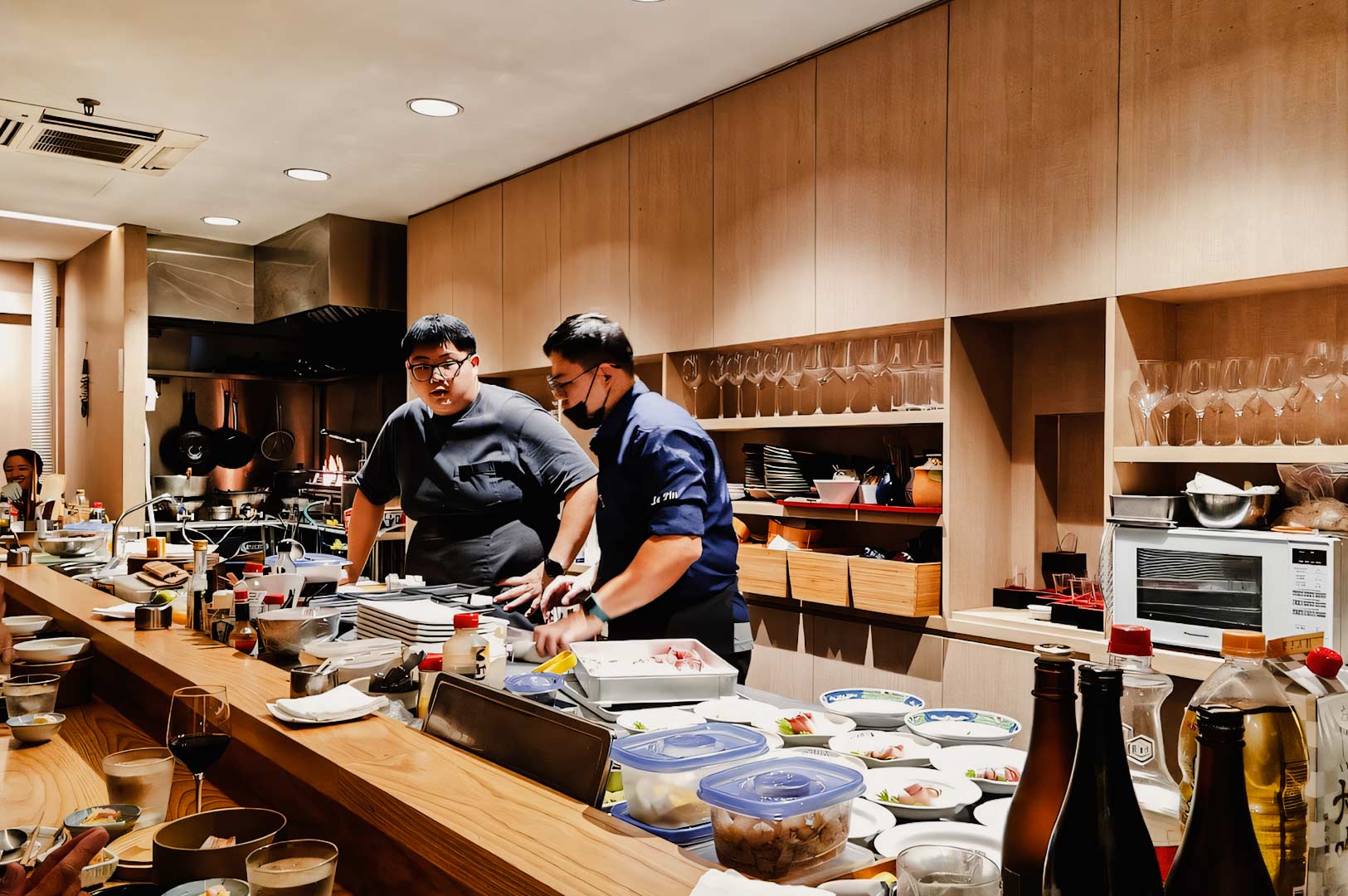 Open-kitchen Japanese dining counter filled with ingredients, cooking tools, and plates as chefs prepare dishes in a modern wood-accented restaurant.