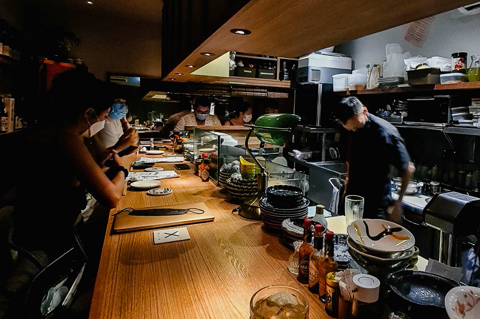Dimly lit open-kitchen restaurant with diners seated at a long wooden counter watching chefs prepare dishes behind a cluttered workstation.