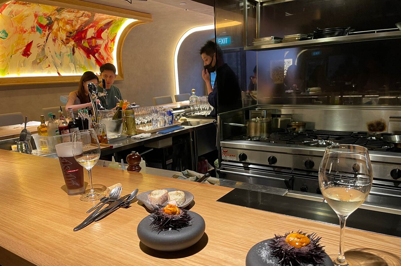 Upscale open-kitchen dining counter with modern lighting, elegant wine glasses, plated uni dishes, and stainless steel cooking equipment in the background.