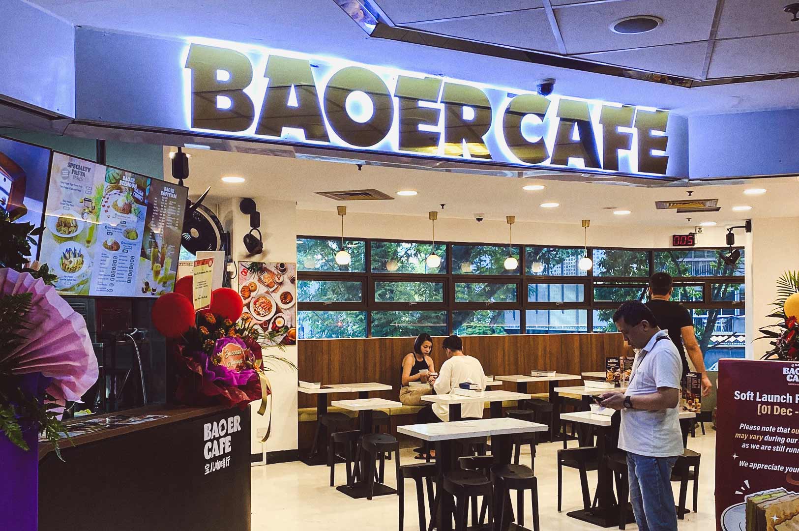 Baoer Cafe interior with modern white walls, digital menu boards, soft launch signage, counter decorated with flowers and balloons, and customers dining.