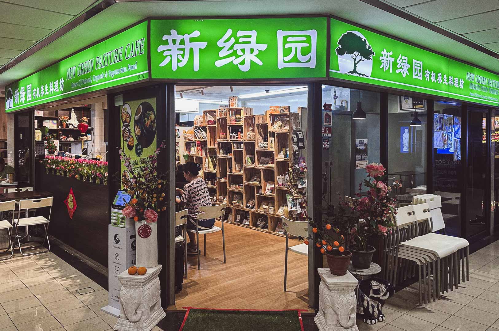 Storefront of New Green Pasture Cafe with bright green sign in English and Chinese, potted plants at entrance, shelves of organic products, and customers seated inside.