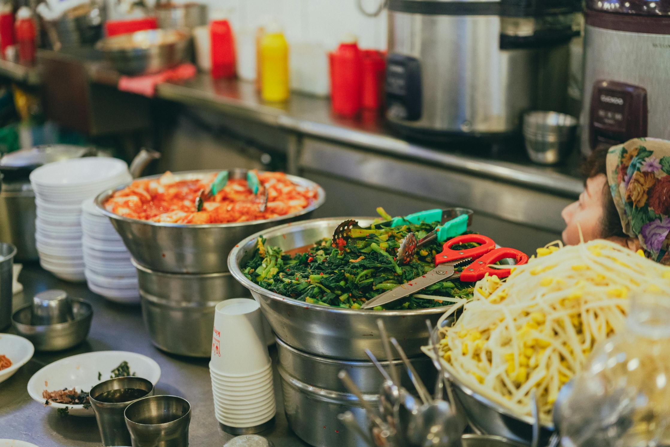 Several large metal bowls filled with colorful Korean side dishes, including kimchi, seasoned greens, and bean sprouts, are lined up on a stainless steel counter in a bustling kitchen. Beside the food, stacks of small white bowls, cups, and kitchen shears are visible, while a person wearing a floral headscarf works in the background.