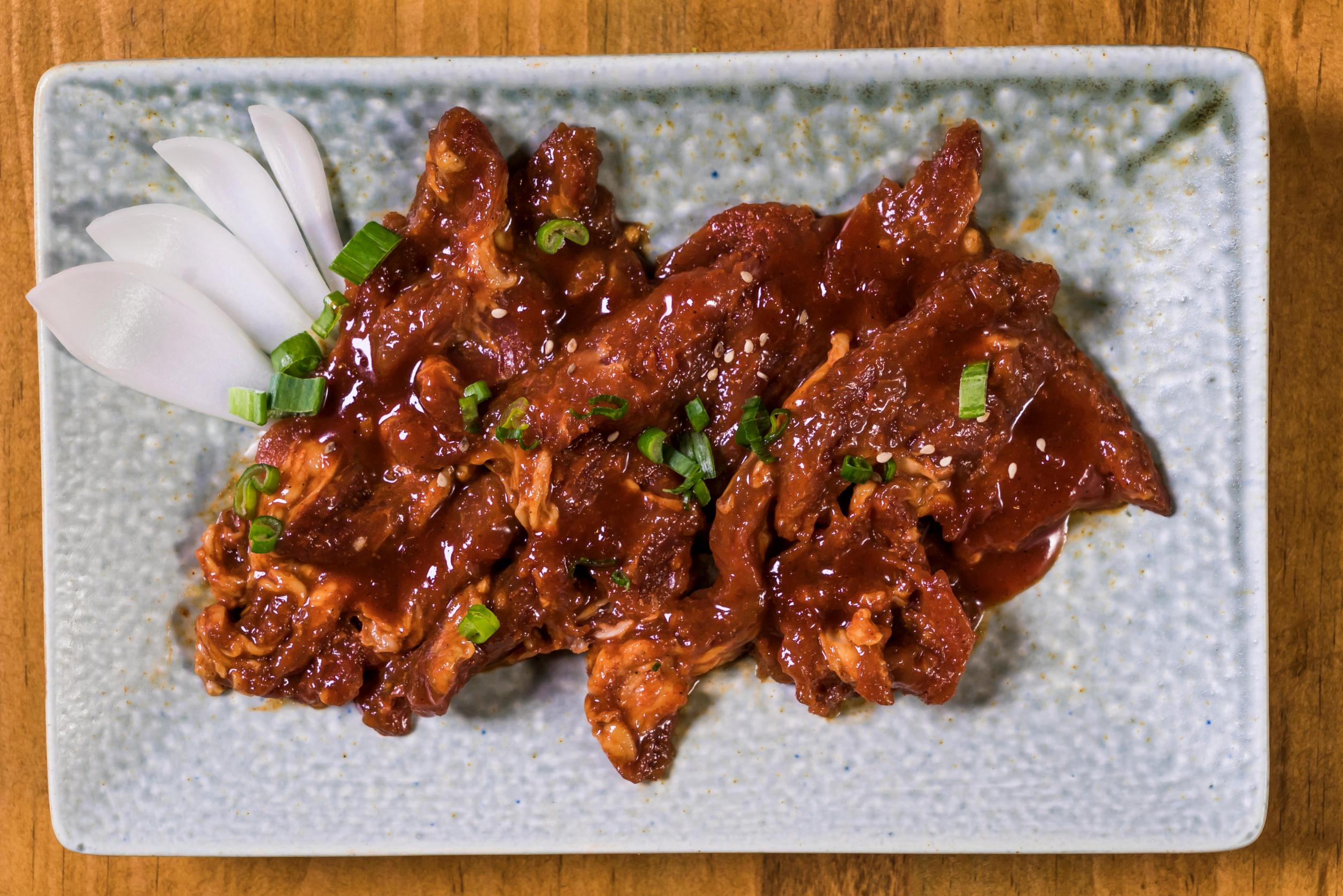 This image displays a rectangular light blue plate topped with thinly sliced meat, likely pork or beef, coated in a dark red, savory glaze. The dish is garnished with a sprinkling of sesame seeds and fresh green onions, with several slices of white onion tucked neatly on the side.