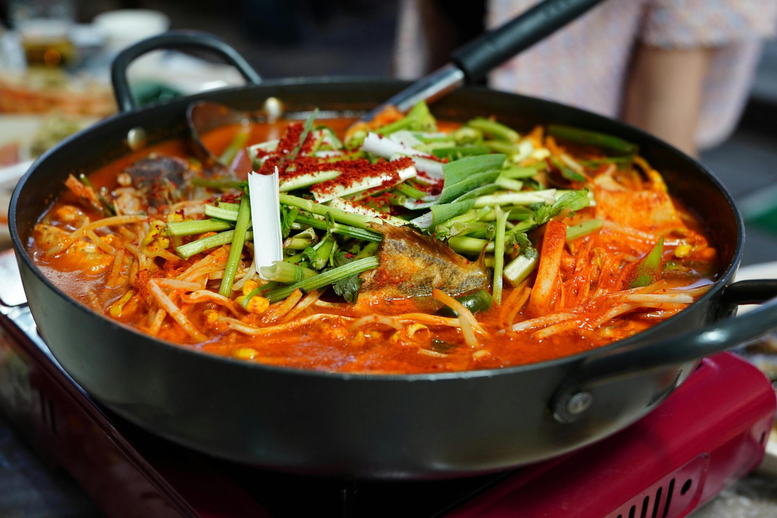 A deep black pot sits on a portable red burner, filled with a bubbling, vibrant red Korean stew containing fish and bean sprouts. The dish is topped with a generous mound of fresh green stalks and a dusting of bright red chili powder.