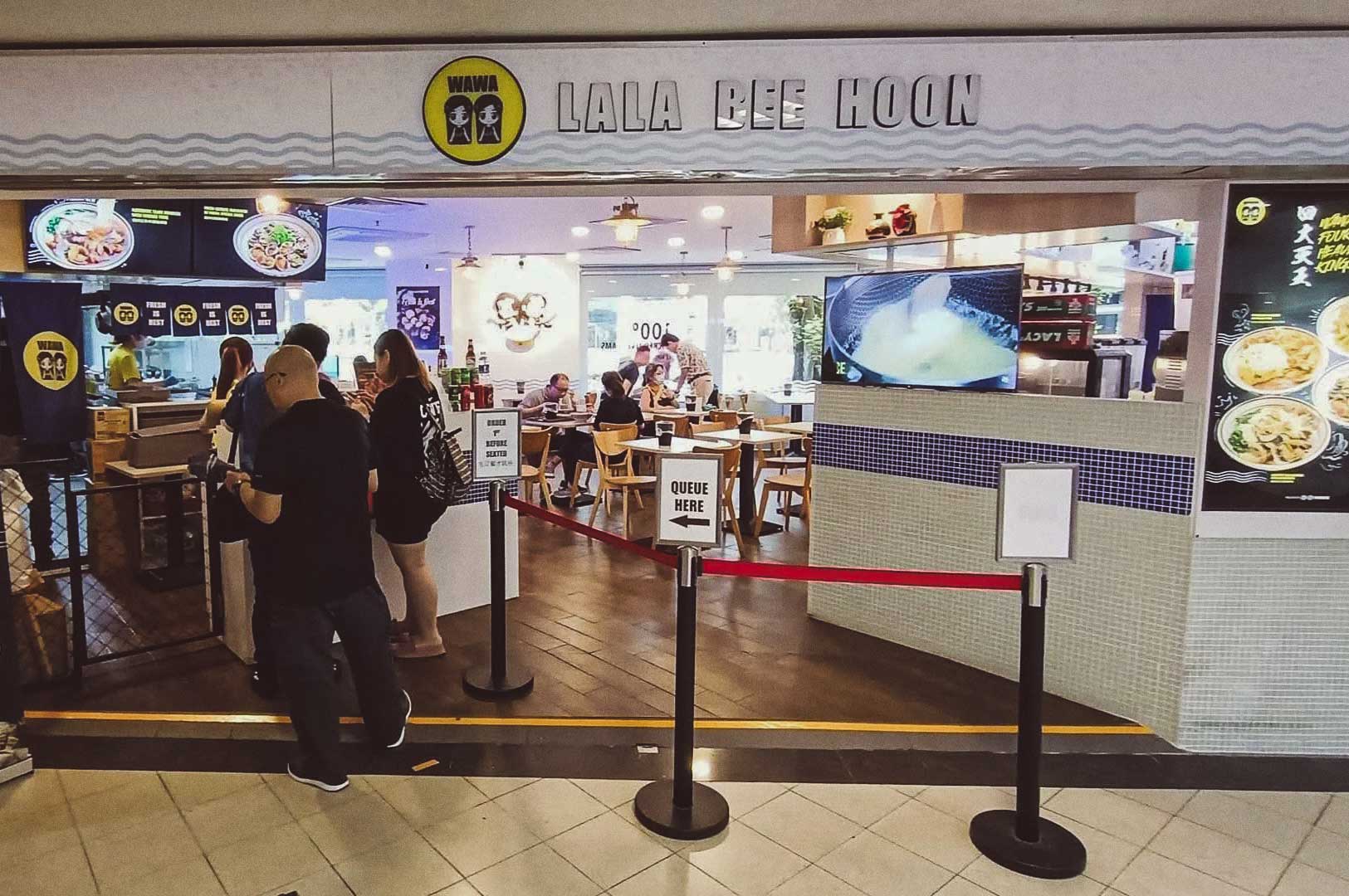 Wawa Lala Bee Hoon restaurant storefront with logo, customer queue, digital menu boards, cooking video screen, and vertical menu display of dishes.