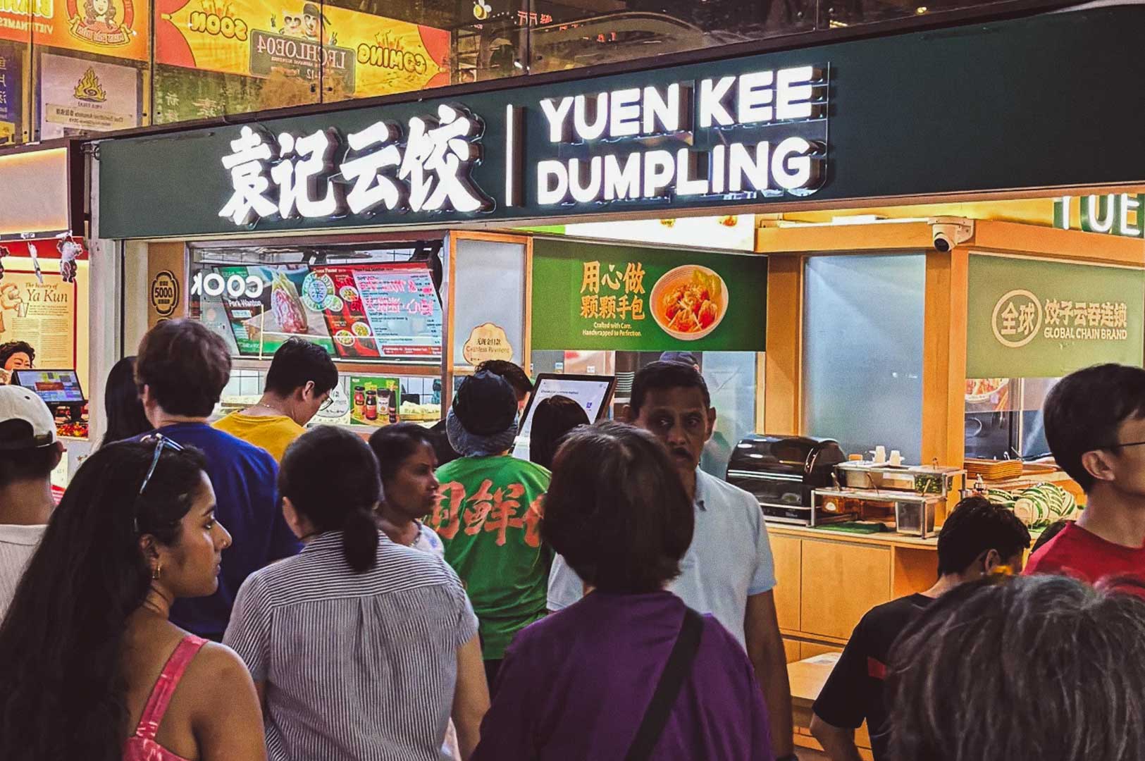 Crowd gathered at Yuen Kee Dumpling stall with bright signs in English and Chinese, menu board, and images of dumplings behind the counter.