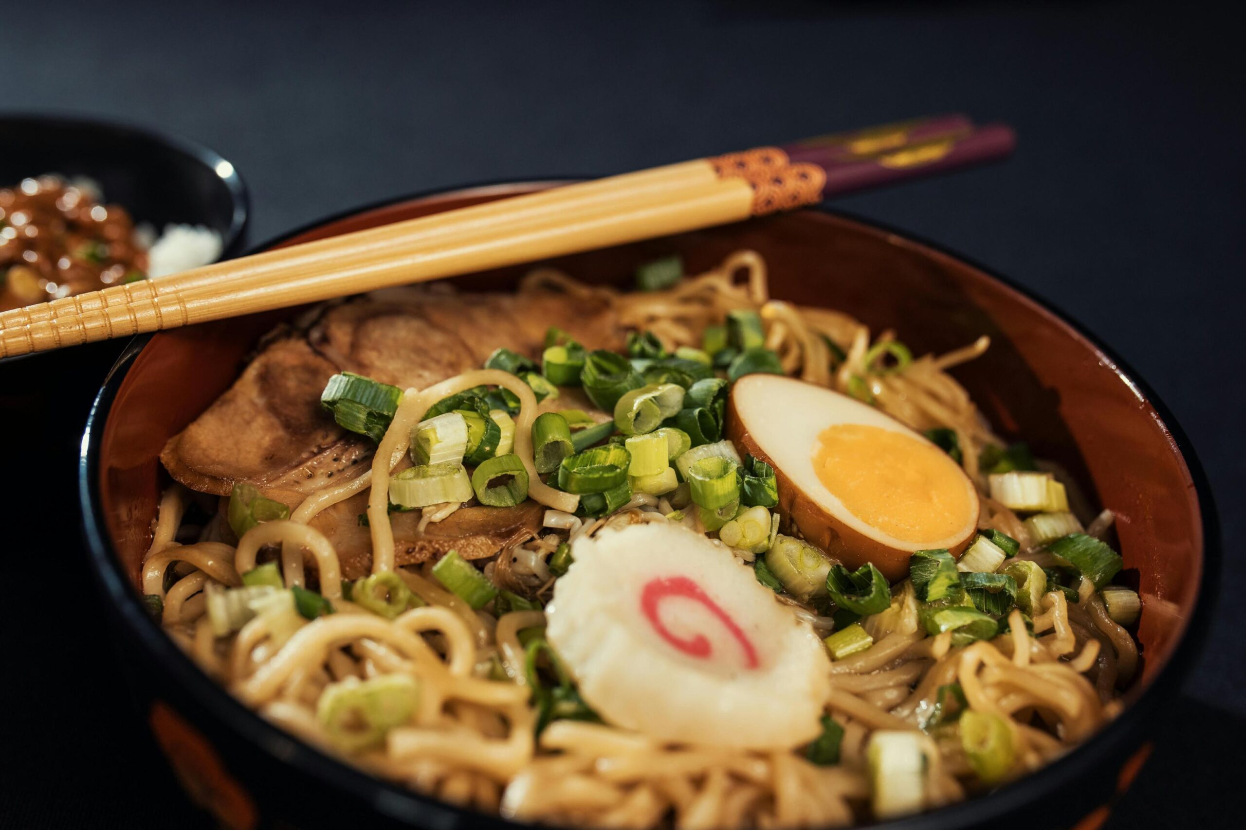 This appetizing close-up features a bowl of noodles generously topped with sliced pork, chopped green onions, a soft-boiled egg, and a slice of pink-swirled fish cake. A pair of wooden chopsticks rests across the rim of the bowl, framing the meal against a dark, moody background.