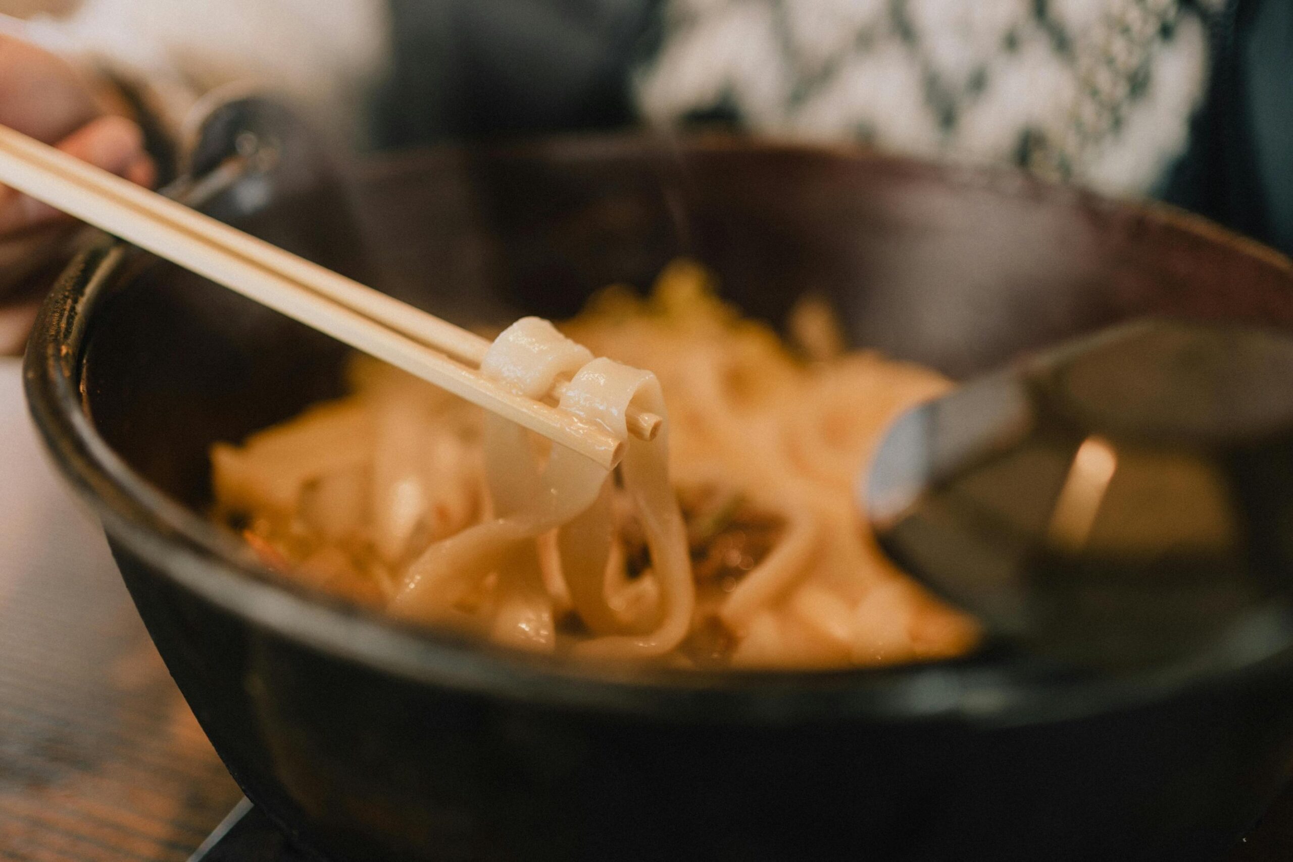 This close-up shot captures a pair of wooden chopsticks lifting thick, glistening noodles from a dark bowl. The shallow depth of field sharply focuses on the texture of the food while softening the background details of the dining environment.