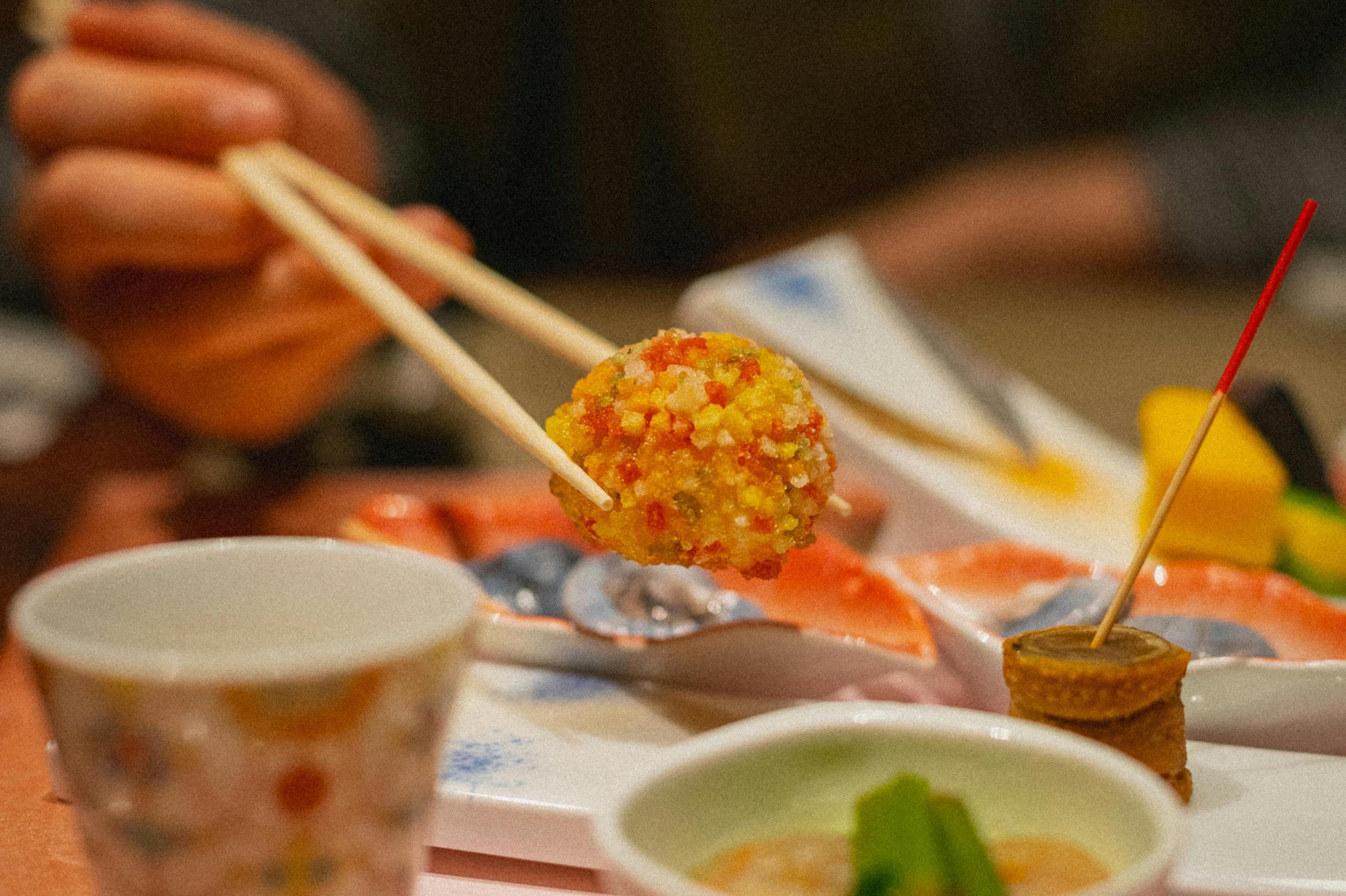 A hand uses wooden chopsticks to lift a round, golden-fried delicacy covered in a colorful, textured coating. The shallow depth of field keeps the food in sharp focus while blurring the surrounding table setting, which features various small plates and a patterned ceramic cup.