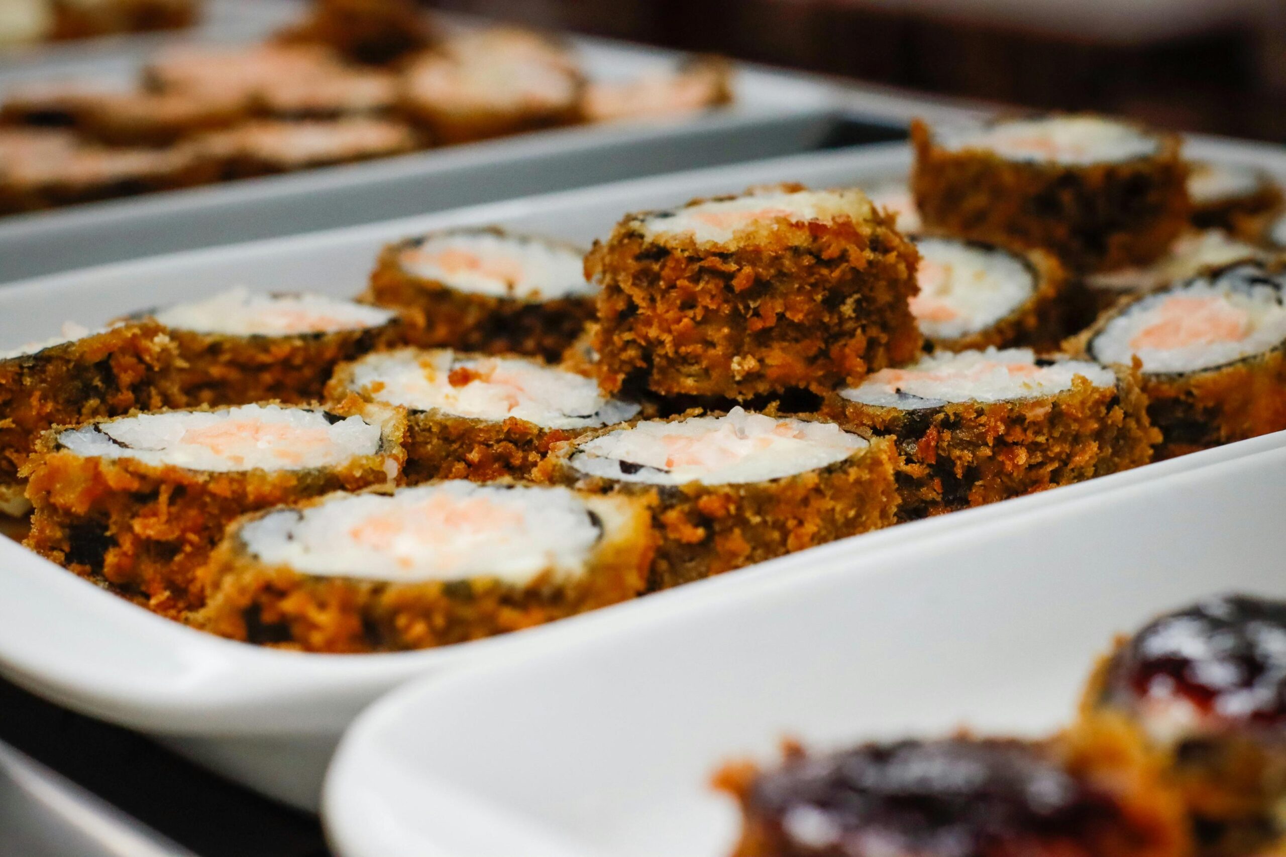A white rectangular platter presents rows of deep-fried sushi rolls, each featuring a crispy golden crust around white rice and a pink filling. One roll rests on top of the others in the center, drawing focus to the textured coating while the surrounding food trays fade into a blur.