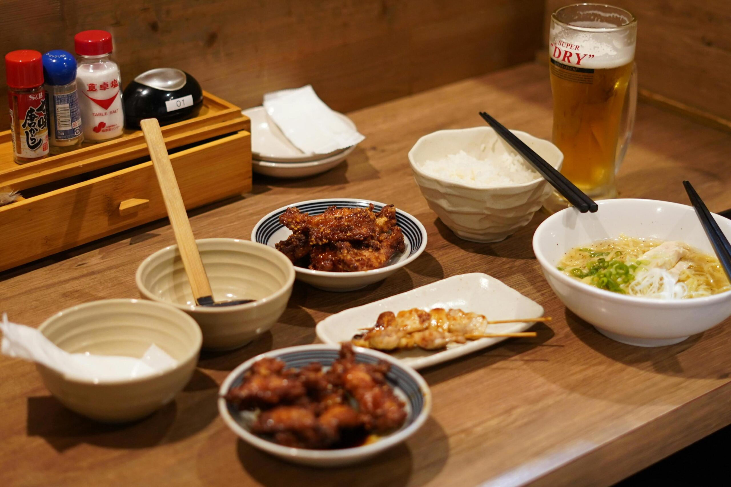 A wooden restaurant table displays a hearty spread including bowls of ramen and white rice, accompanied by small plates of fried chicken and skewers. A tall glass of beer stands next to condiment jars and empty bowls, completing the casual dining setup.