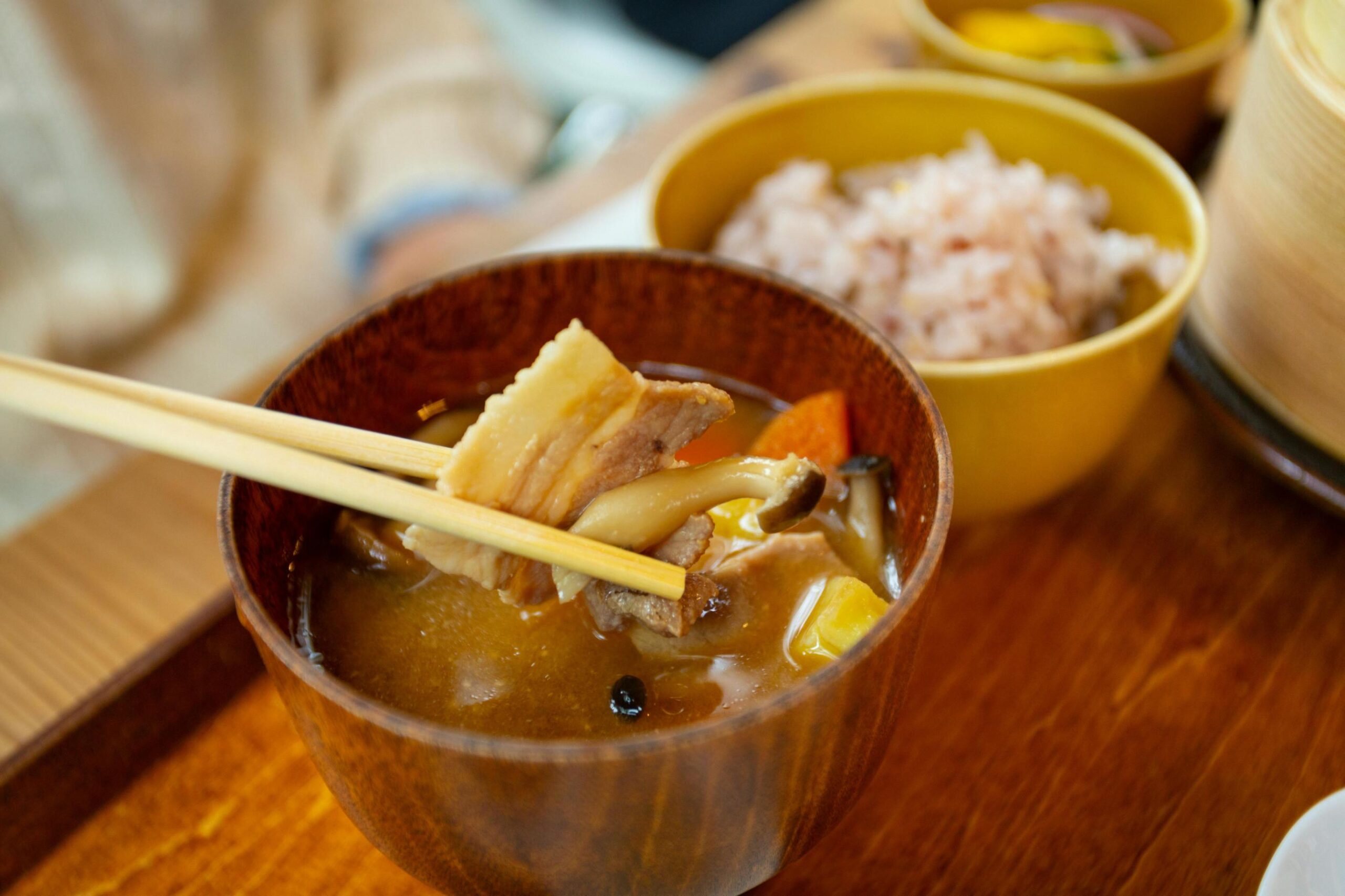 Wooden chopsticks are captured lifting a slice of pork belly and a mushroom from a dark wooden bowl filled with savory soup and vegetables. A yellow bowl of rice sits in the blurred background, rounding out this warm and comforting meal set on a wooden tray.