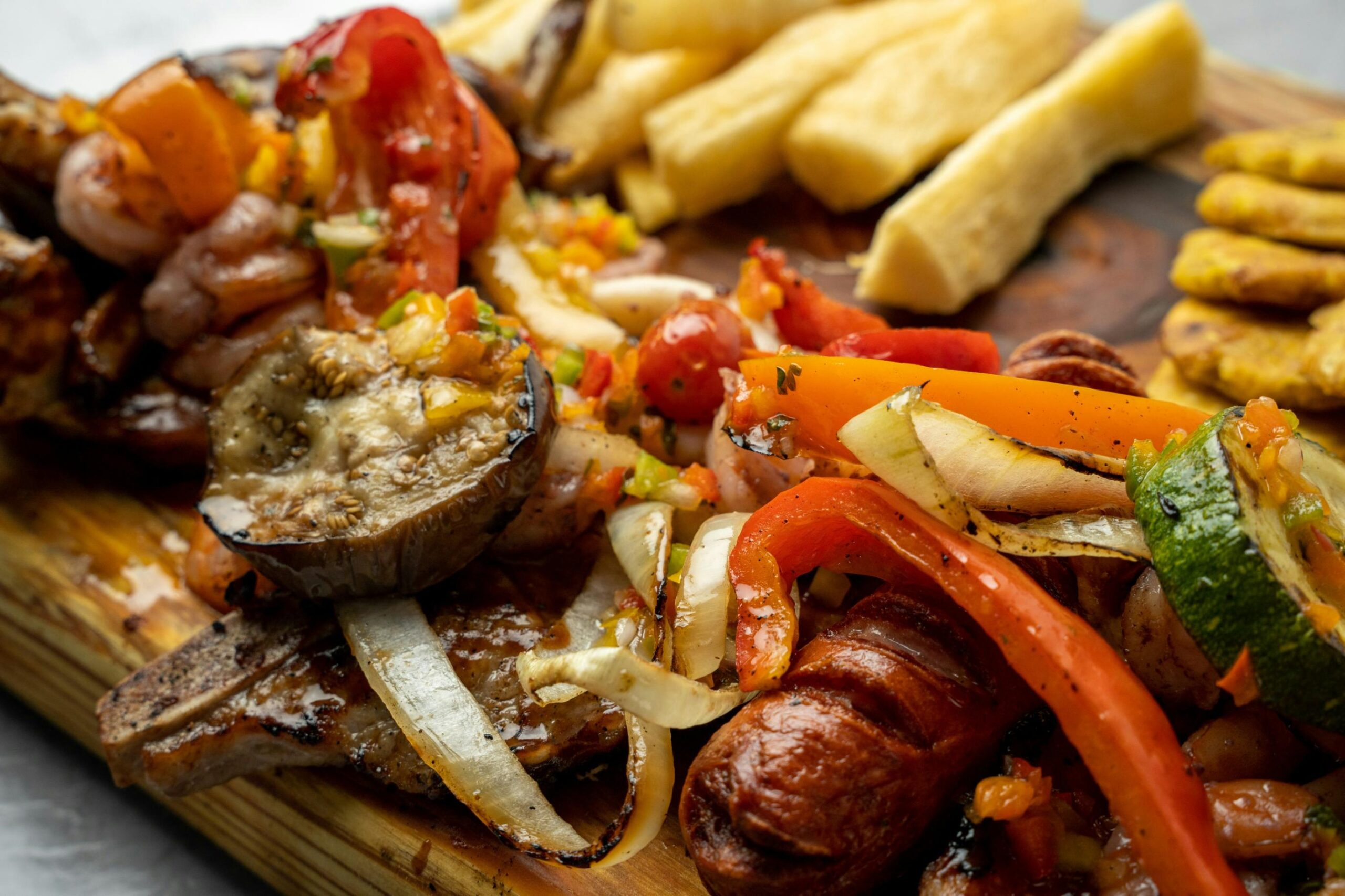 A vibrant wooden platter is piled high with a variety of grilled foods, including charred sausages, steaks, and shrimp mixed with colorful peppers and onions. To the side, thick-cut yuca fries and golden-brown plantains complete this hearty, rustic meal.