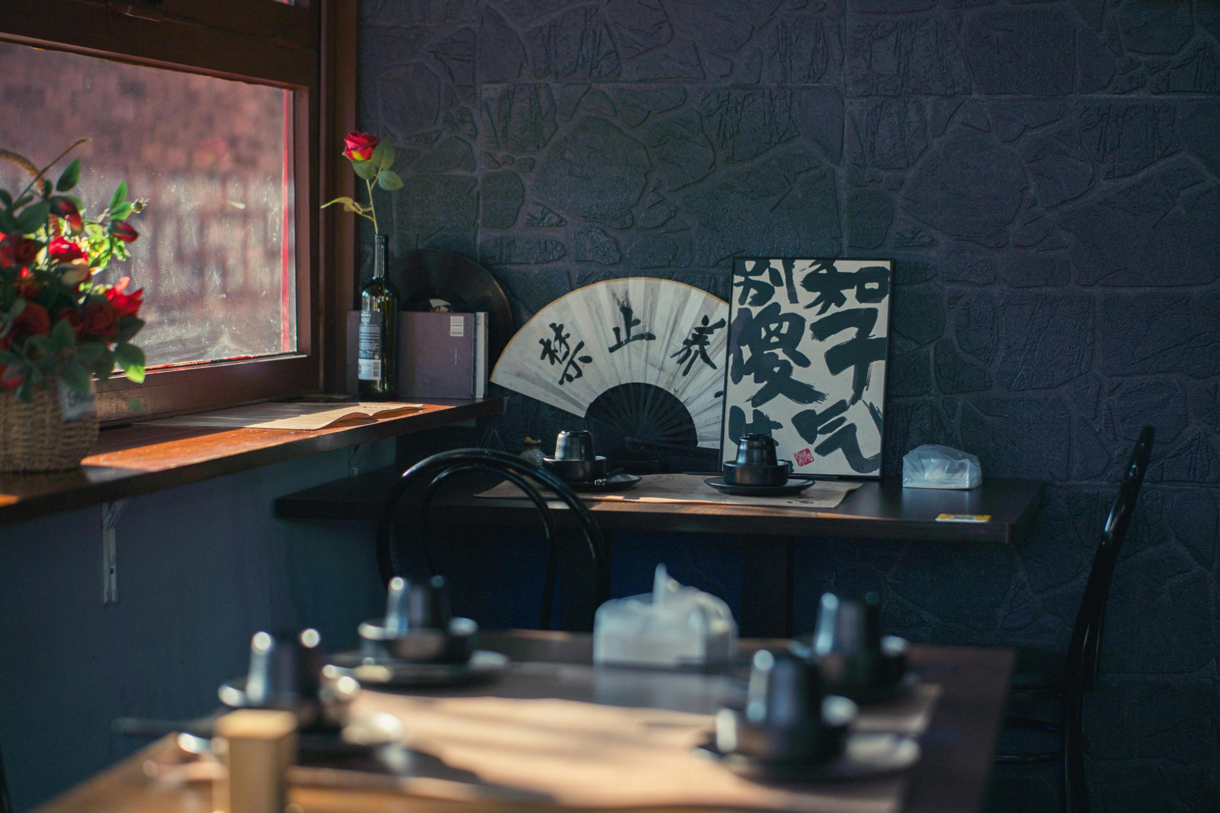This cozy restaurant interior features dark stone-textured walls adorned with a decorative folding fan and calligraphy art. Natural light streams through a window onto wooden tables set with dark ceramic teaware, creating a serene and inviting atmosphere.