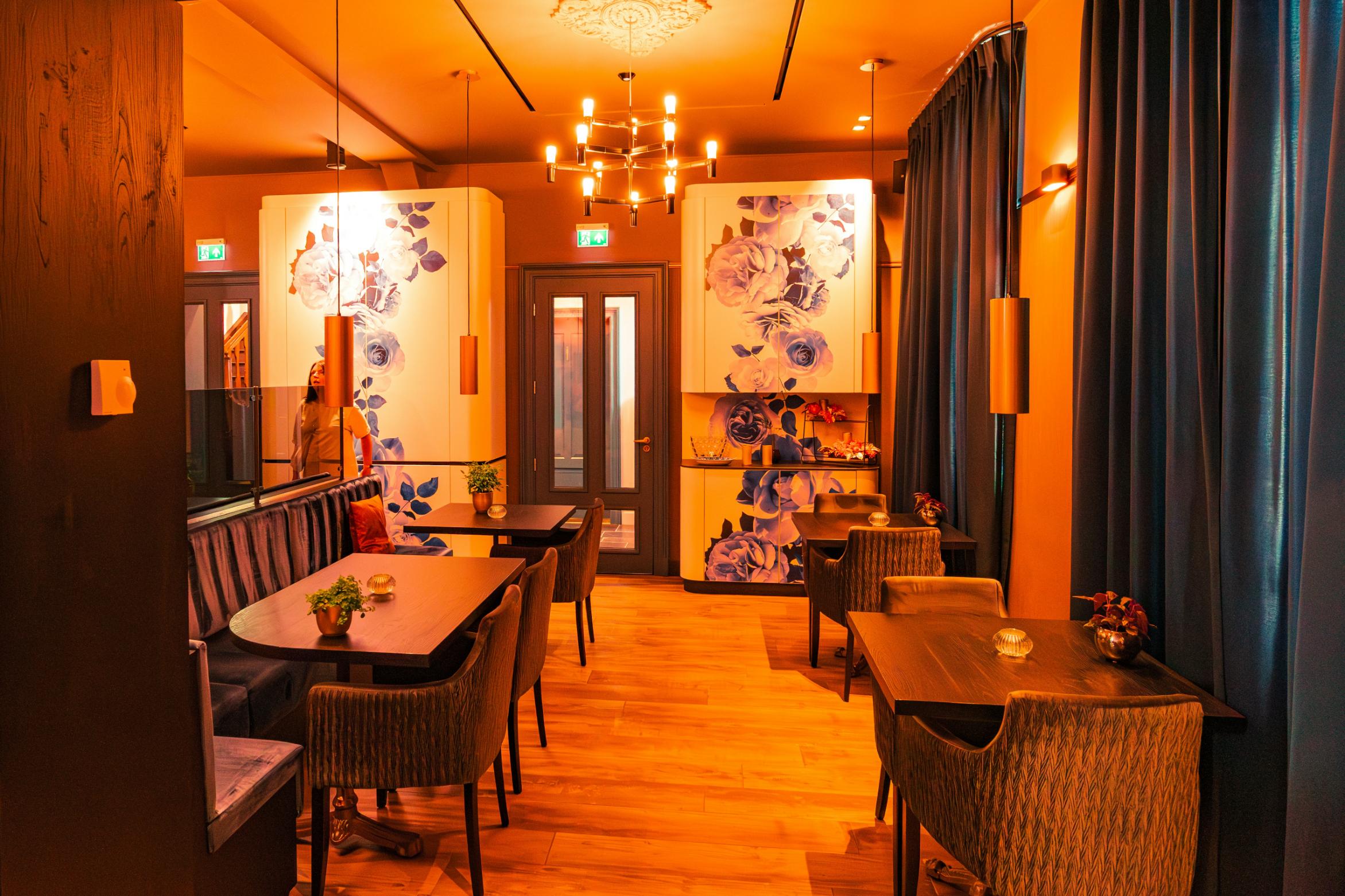 This elegant dining room is bathed in warm, golden light and features striking blue floral mural cabinets that serve as focal points. Dark wood tables and plush velvet seating are complemented by long blue curtains and a modern multi-bulb chandelier, creating a sophisticated and cozy ambiance.