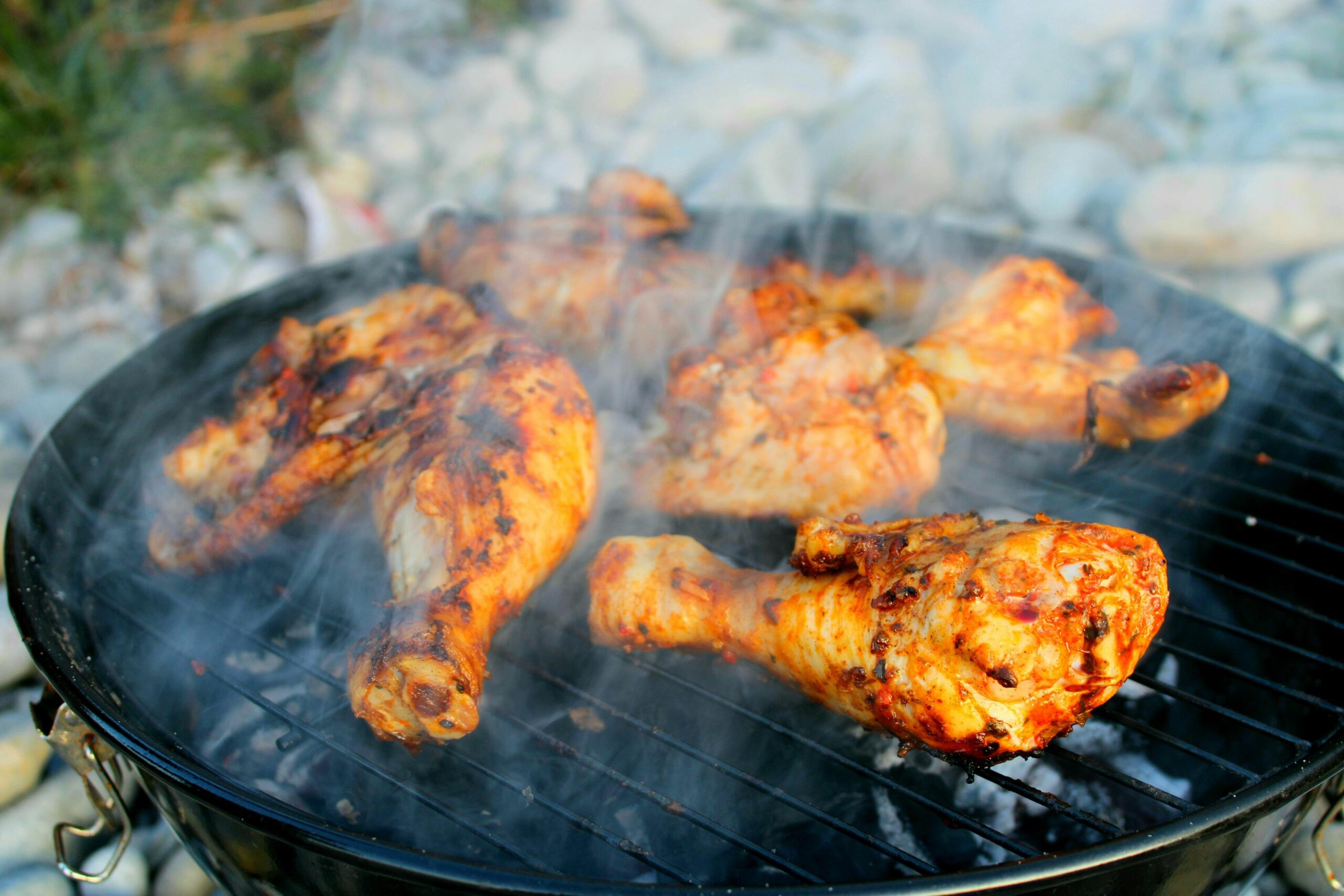 Several pieces of seasoned chicken legs and thighs sizzle over a charcoal grill, releasing plumes of white smoke as they develop a charred, golden-brown crust. The outdoor setup is positioned against a background of light-colored river stones, capturing a rustic and flavorful barbecue scene.