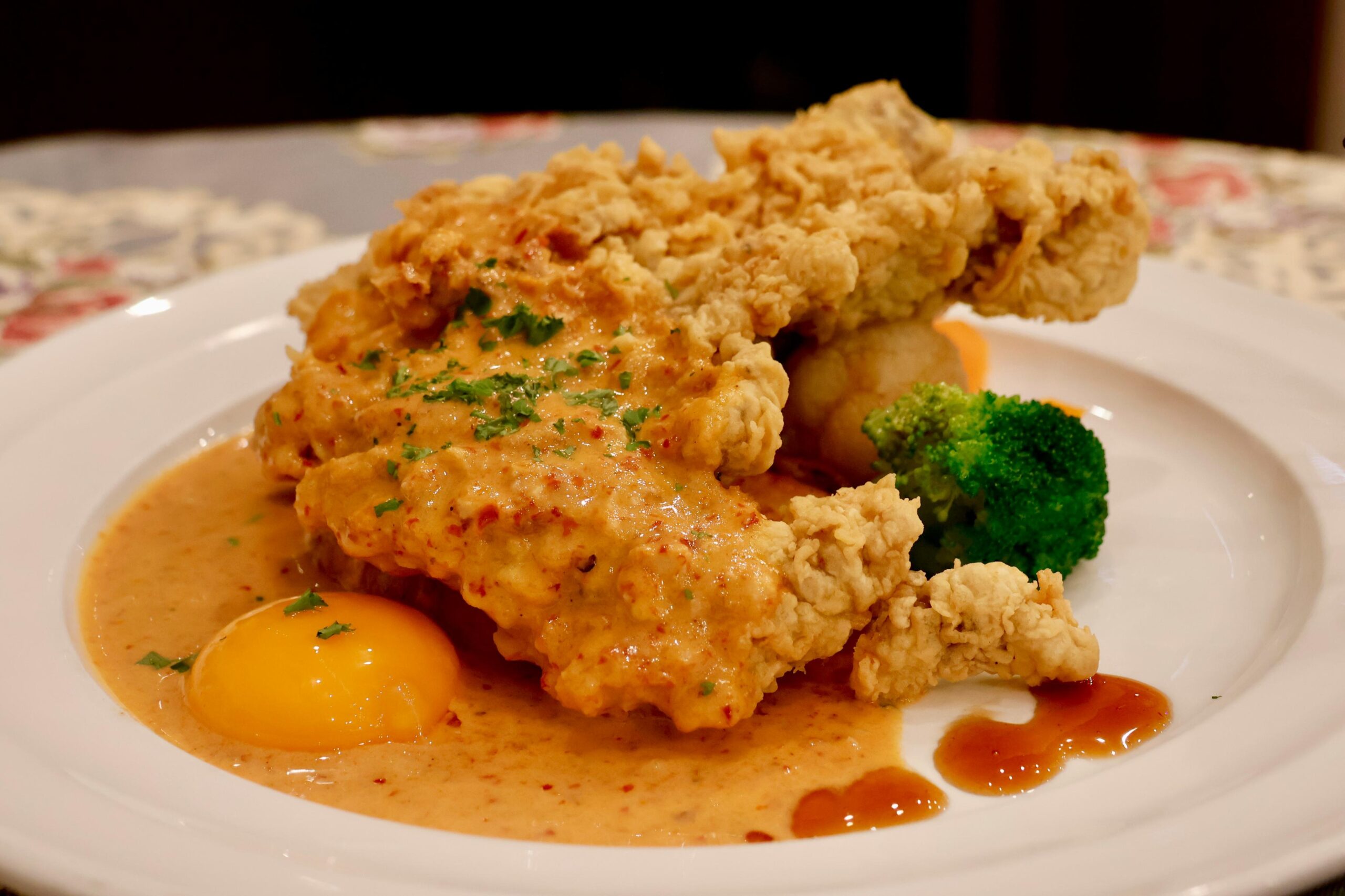 A crispy, golden-brown piece of fried chicken is served on a white plate, partially covered in a rich, creamy orange sauce sprinkled with herbs. A bright yellow egg yolk sits nestled in the sauce alongside a small floret of steamed broccoli, creating a visually indulgent and unique dish.
