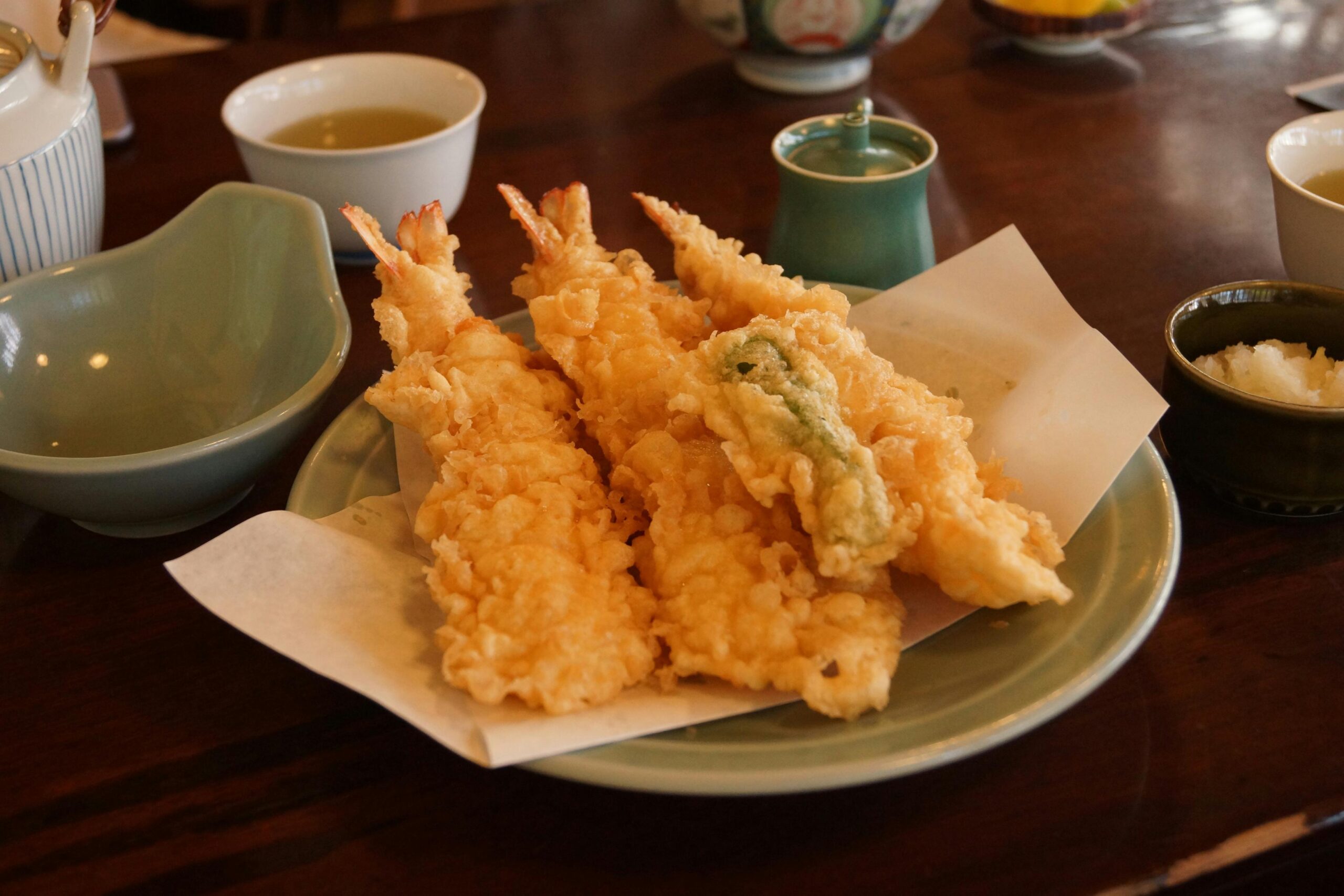 This appetizing close-up features several pieces of crispy, golden-brown shrimp and vegetable tempura served on a sheet of white paper atop a pale green plate. The dish rests on a dark wooden table accompanied by traditional dining elements, including a small bowl of grated daikon radish, a ceramic sauce container, and a cup of tea.