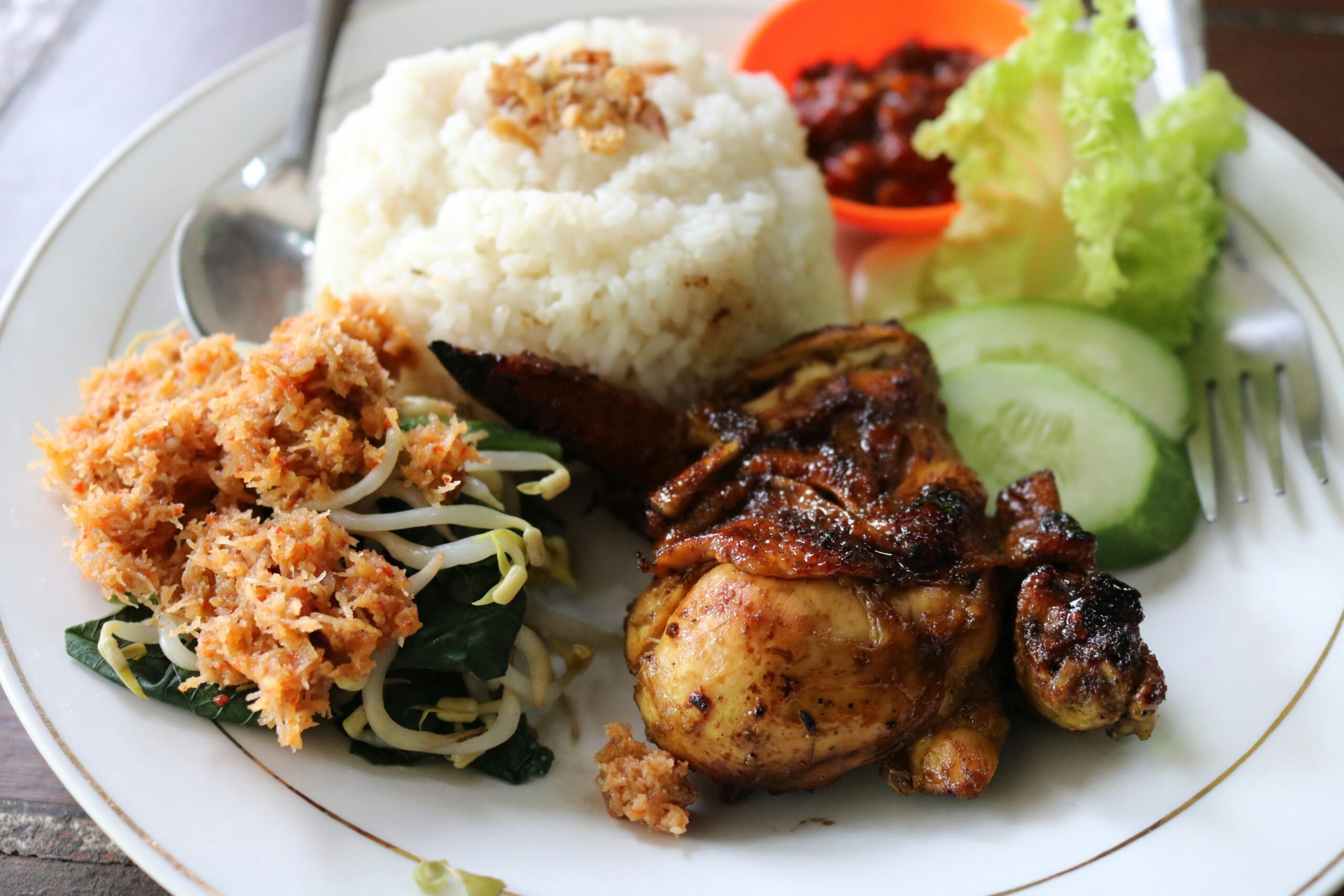This appetizing meal features a mound of white rice topped with crispy fried shallots, served alongside a piece of dark, glazed grilled chicken and a portion of vegetables mixed with spiced grated coconut. Fresh cucumber slices and lettuce garnish the white plate, which also holds a small orange bowl of red chili paste to complete the dish.