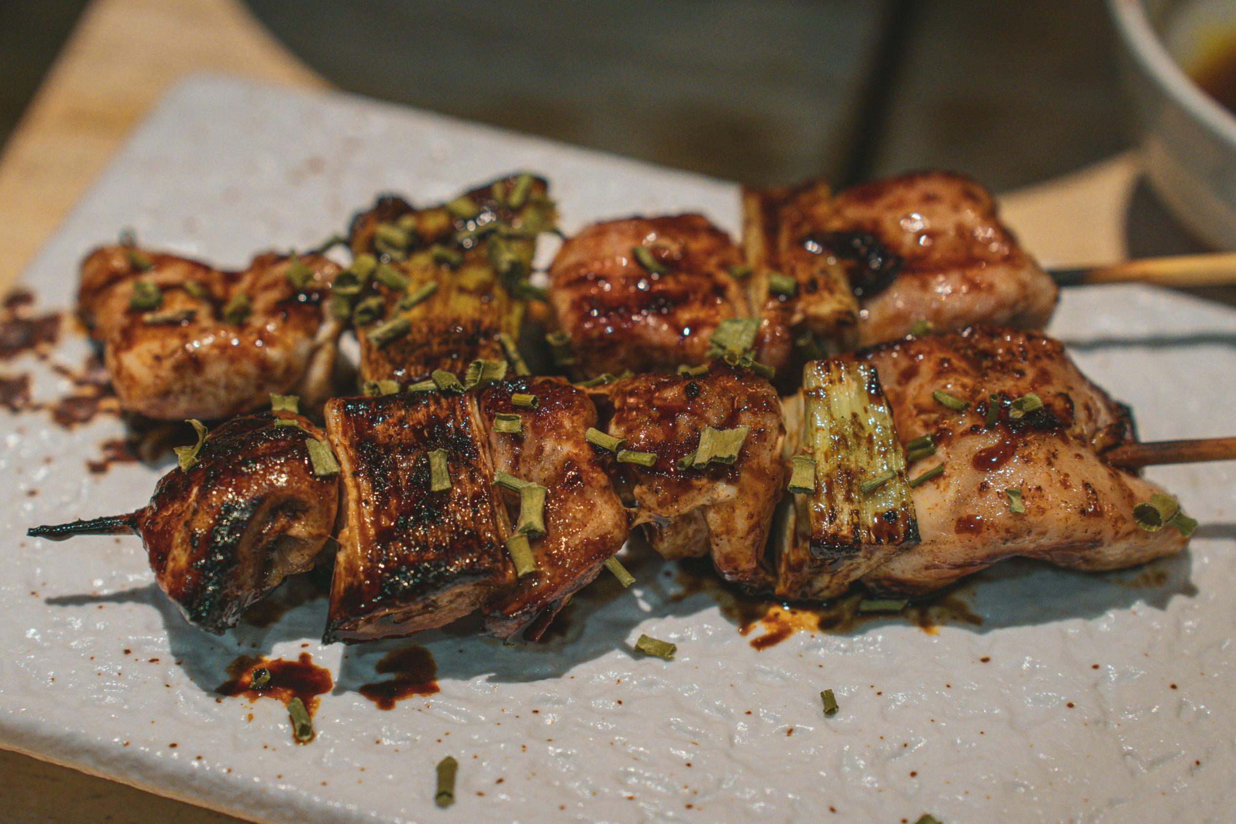 This close-up captures two succulent skewers of grilled meat and leeks, likely yakitori, glazed in a rich, dark sauce and sprinkled with chopped green herbs. The skewers rest on a textured white plate, displaying appetizing char marks that suggest a smoky, savory flavor profile.