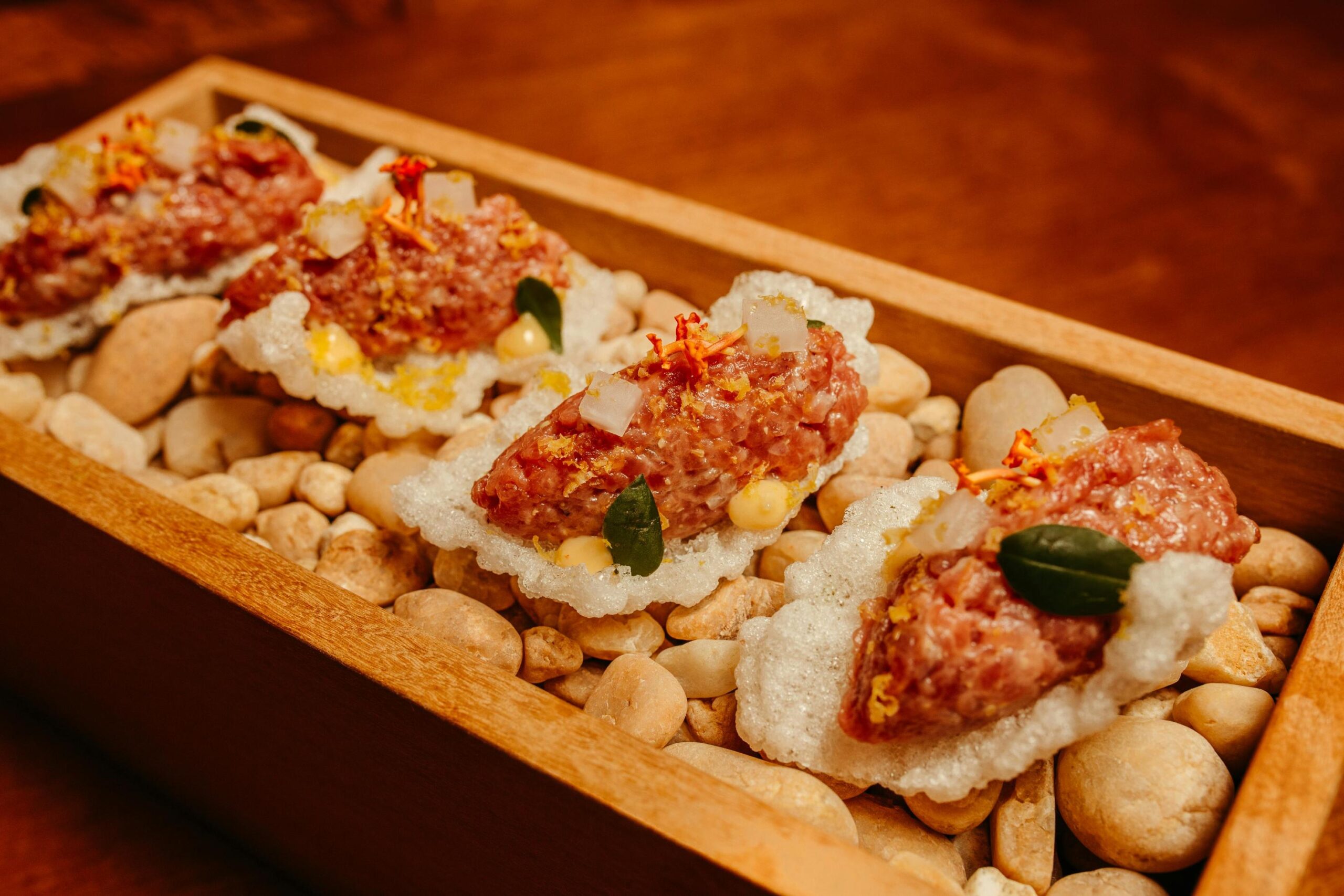 This elegant culinary display features four portions of seasoned raw meat, likely beef tartare, served on crispy white crackers. Arranged in a row within a wooden box filled with smooth river stones, each bite is intricately topped with small white cubes, fresh green leaves, and delicate orange garnishes.