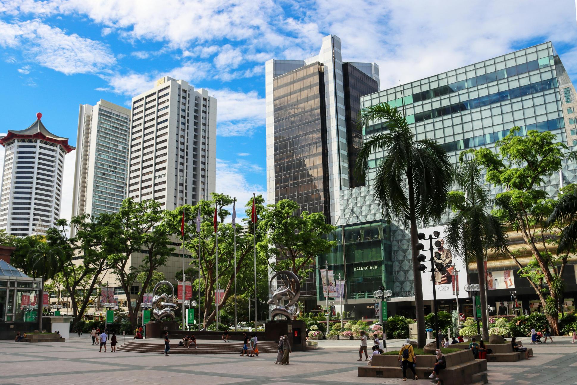This vibrant urban plaza features a spacious paved area filled with pedestrians and scattered palm trees, set against a backdrop of towering modern skyscrapers under a bright blue sky. The scene is anchored by distinctive architecture, including a building topped with a traditional red pagoda roof and abstract metal sculptures located near a luxury retail storefront.