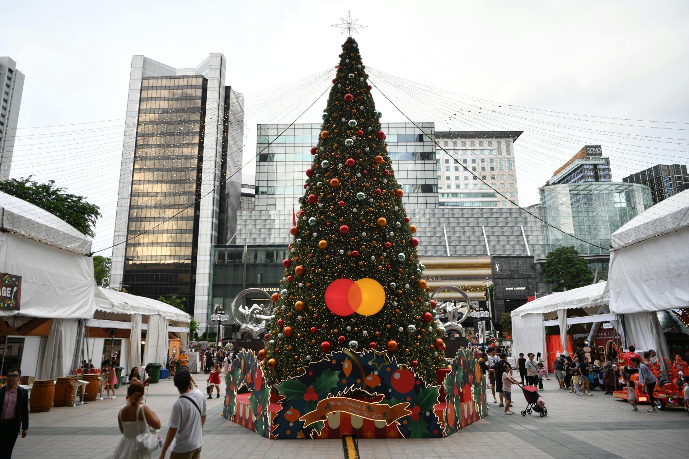 This festive urban scene centers on a towering Christmas tree decorated with colorful ornaments and a prominent Mastercard logo, standing in the middle of a paved plaza. Surrounded by white event tents and pedestrians, the holiday display is set against a backdrop of modern glass skyscrapers under an overcast sky.