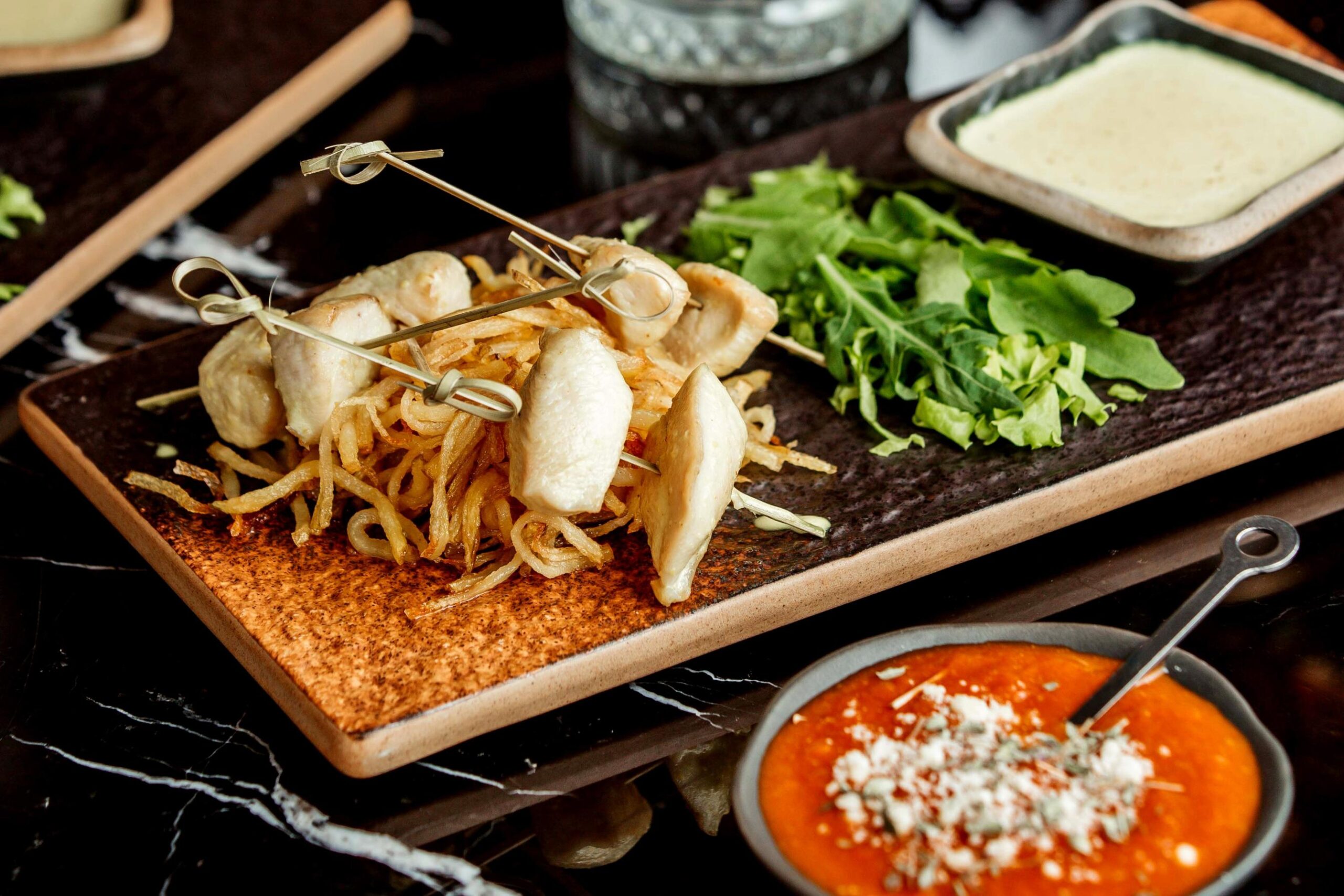Skewered chicken fillet pieces are presented atop a mound of crispy shoestring fries, accompanied by a side of fresh arugula on a rustic rectangular plate. Two dipping sauces, one creamy and one vibrant red with garnish, complete the appetizing display set against a dark marble surface.