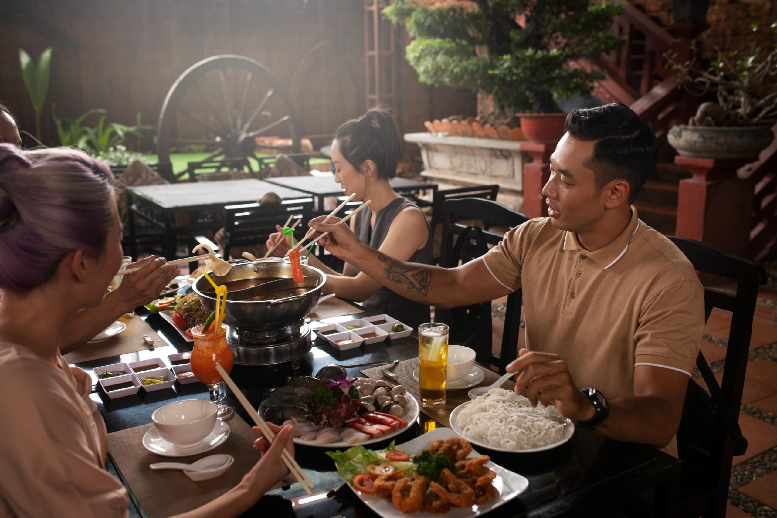 A group of friends enjoys a communal hot pot meal at a rustic outdoor restaurant, using chopsticks to dip ingredients into the central steaming vessel. The table is crowded with plates of raw seafood, noodles, and fried dishes, surrounded by traditional decor such as a large wooden wheel and potted plants.