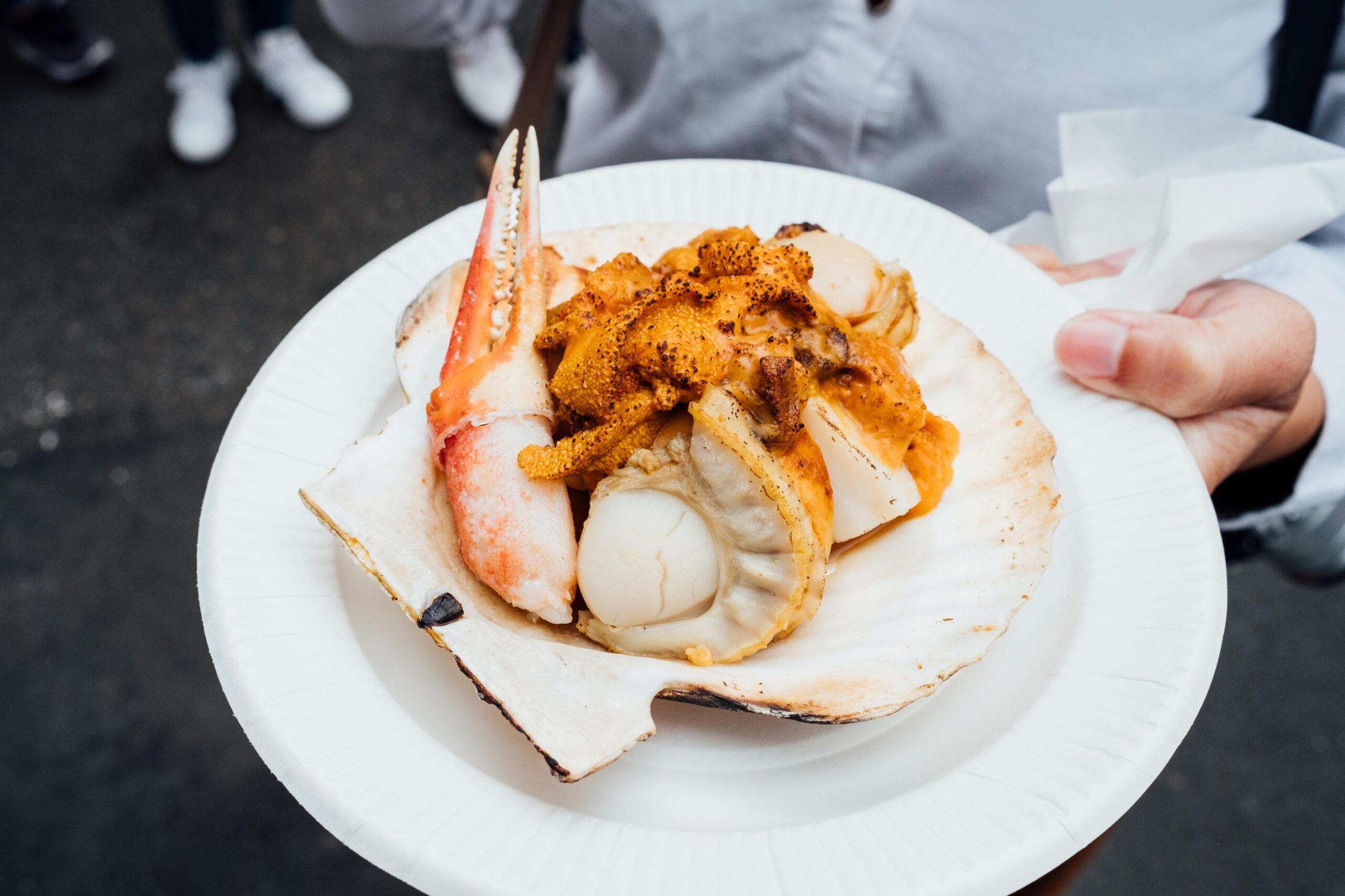 A hand holds a white paper plate featuring a large grilled scallop shell filled with an assortment of seafood, including a prominent crab claw and plump scallop meat. The dish is topped with a rich, torched orange garnish and is set against a blurred background of pavement and bystanders, evoking a bustling street food atmosphere.