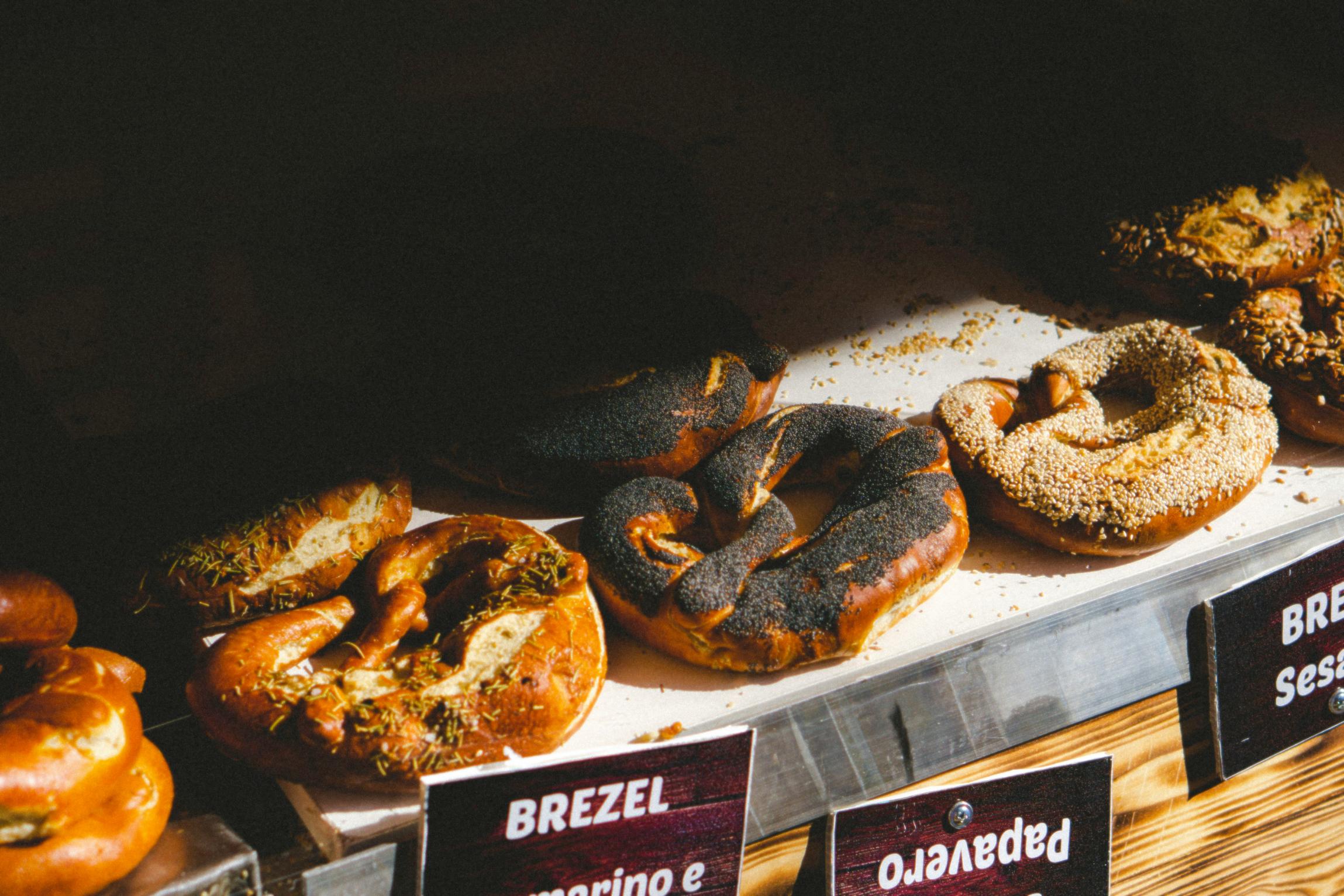 A bakery display features a row of golden-brown soft pretzels topped with ingredients like poppy seeds, sesame seeds, and herbs. Bright natural light casts a spotlight on the textured pastries and their labels, contrasting sharply with the deep shadows behind them.