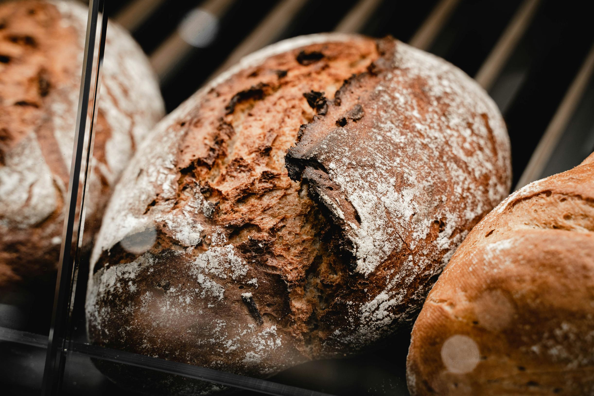 A close-up shot reveals a rustic loaf of artisan bread with a dark, crusty exterior that has been lightly dusted with flour. The central loaf is in sharp focus, highlighting the deep, jagged cracks and rich texture of its surface, while adjacent loaves fade slightly into the background.