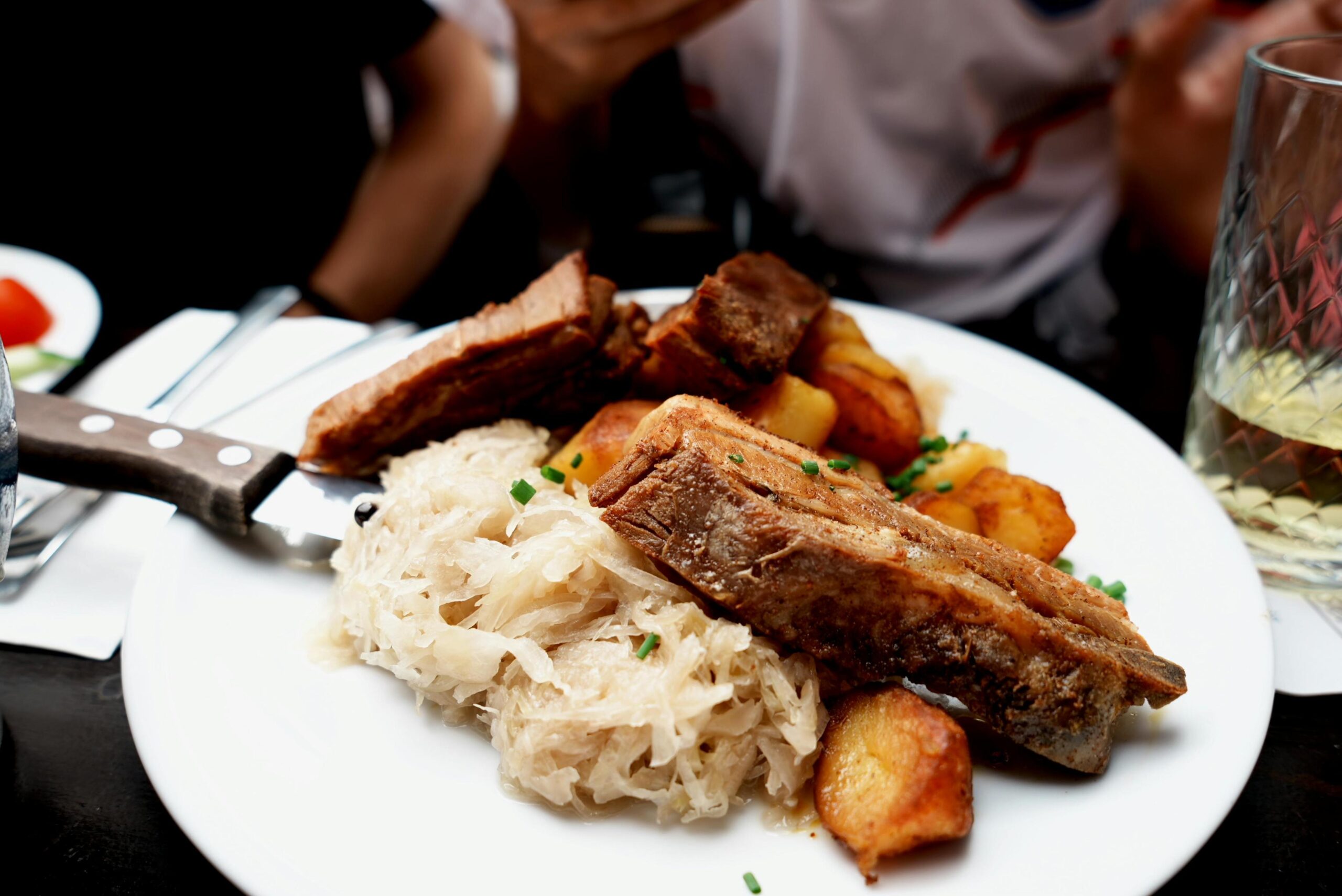 A hearty plate of food features chunks of crispy roasted pork served alongside golden potatoes and a mound of sauerkraut. Fresh chopped chives are sprinkled over the meal, which is set on a table with dining utensils and a drinking glass visible nearby.