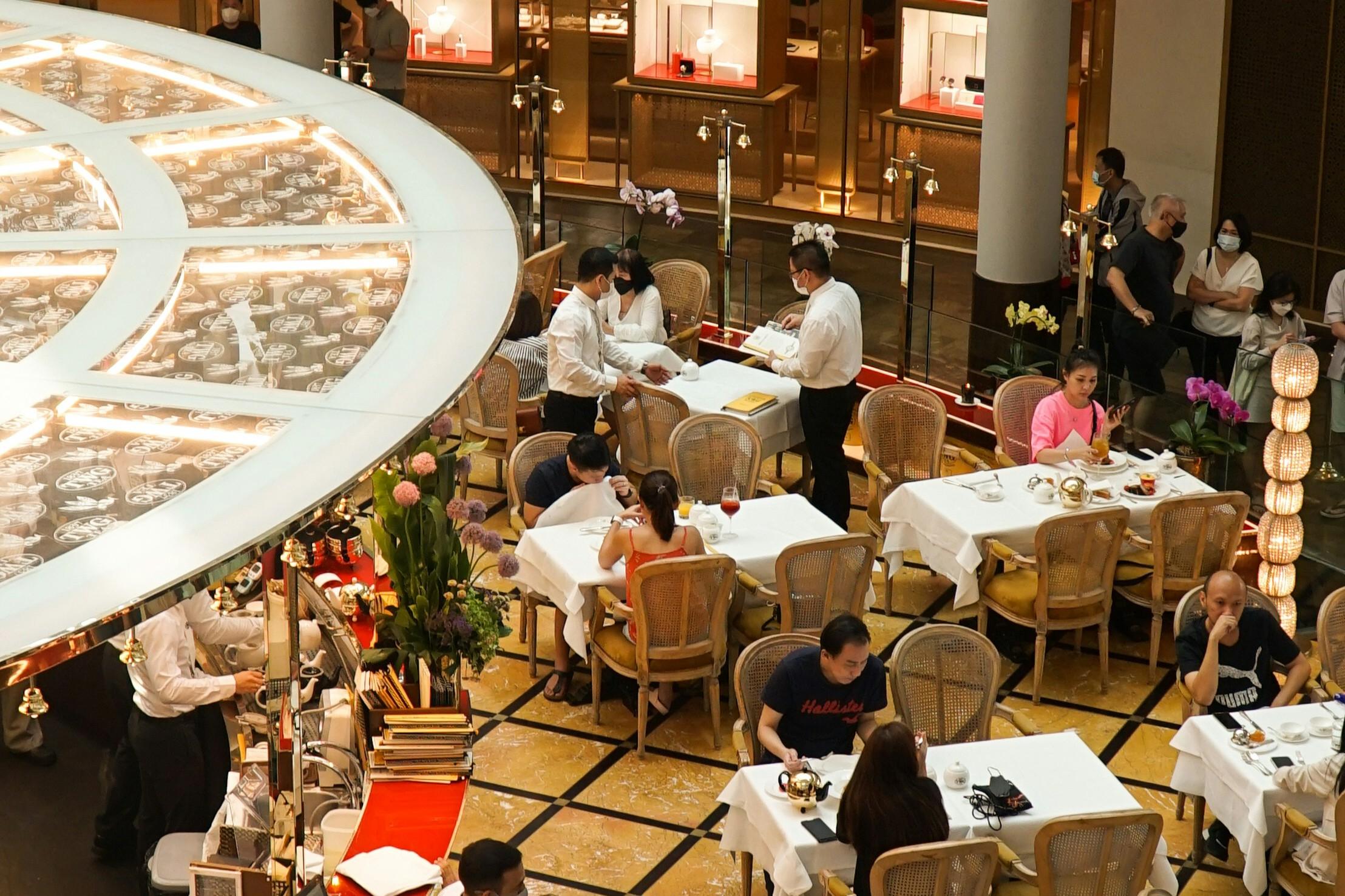 SG Malls Dining Guide - 3 This upscale dining venue features white-clothed tables and elegant rattan chairs arranged on a patterned yellow and black floor. Uniformed waitstaff attend to seated patrons near a curved service counter, which is framed by a distinctive illuminated overhead fixture and tall floral arrangements.