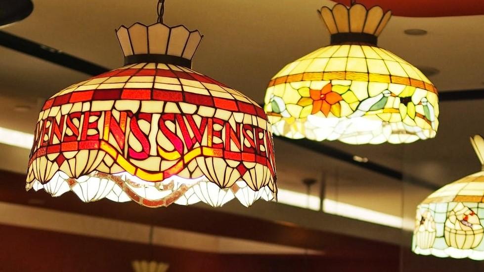 This image captures a close-up of illuminated Tiffany-style stained glass lamps hanging from a ceiling, with the foreground fixture prominently displaying the text "SWENSENS" in red lettering. Behind it, other colorful lamps feature floral or decorative patterns, contributing to a warm, vintage atmosphere.