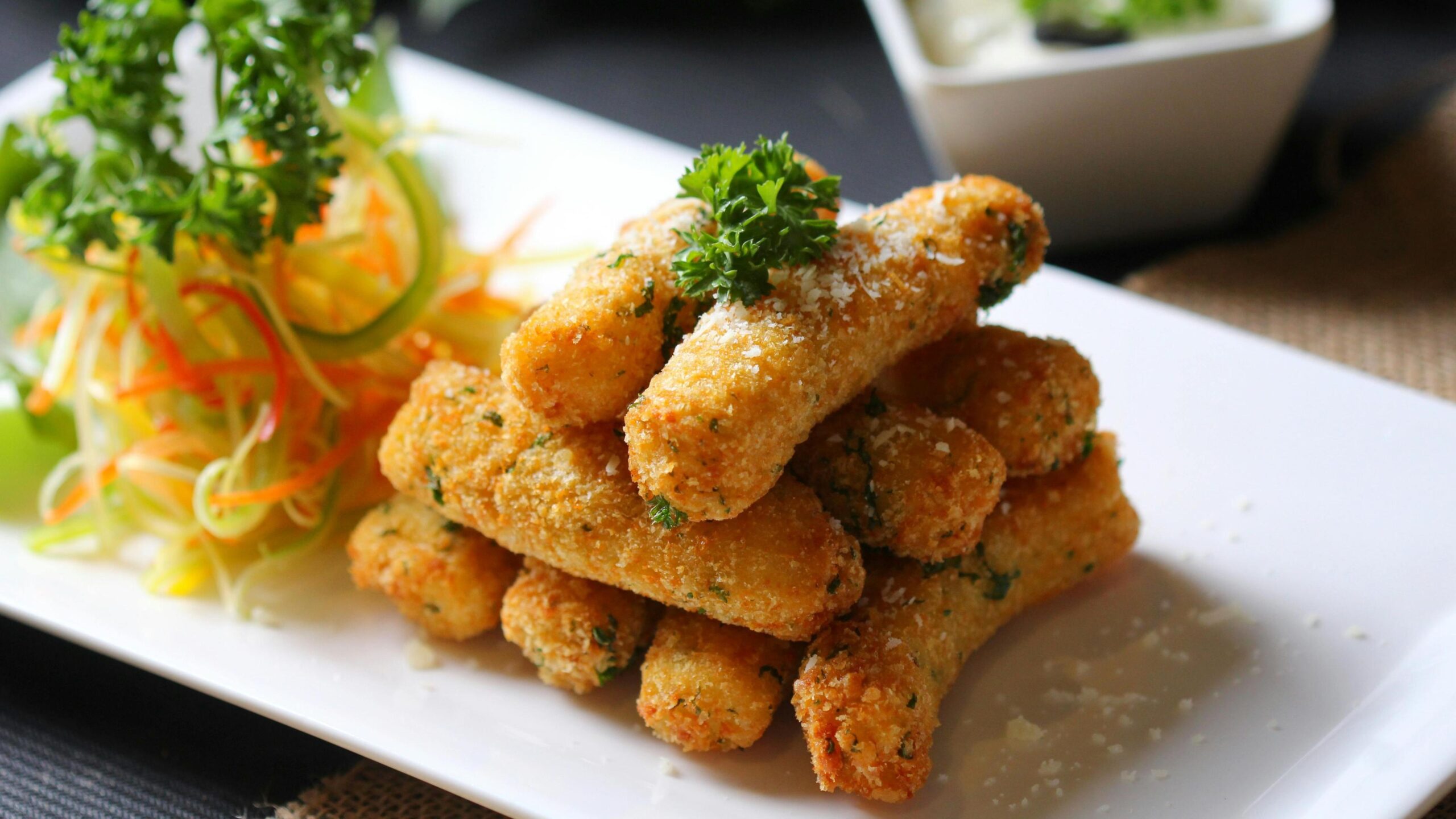 A stack of crispy, golden-brown fried sticks is elegantly arranged on a white rectangular plate, garnished with fresh parsley and a light dusting of grated cheese. To the left, a colorful side salad of shredded vegetables adds contrast, while a small bowl of creamy dipping sauce sits in the background.