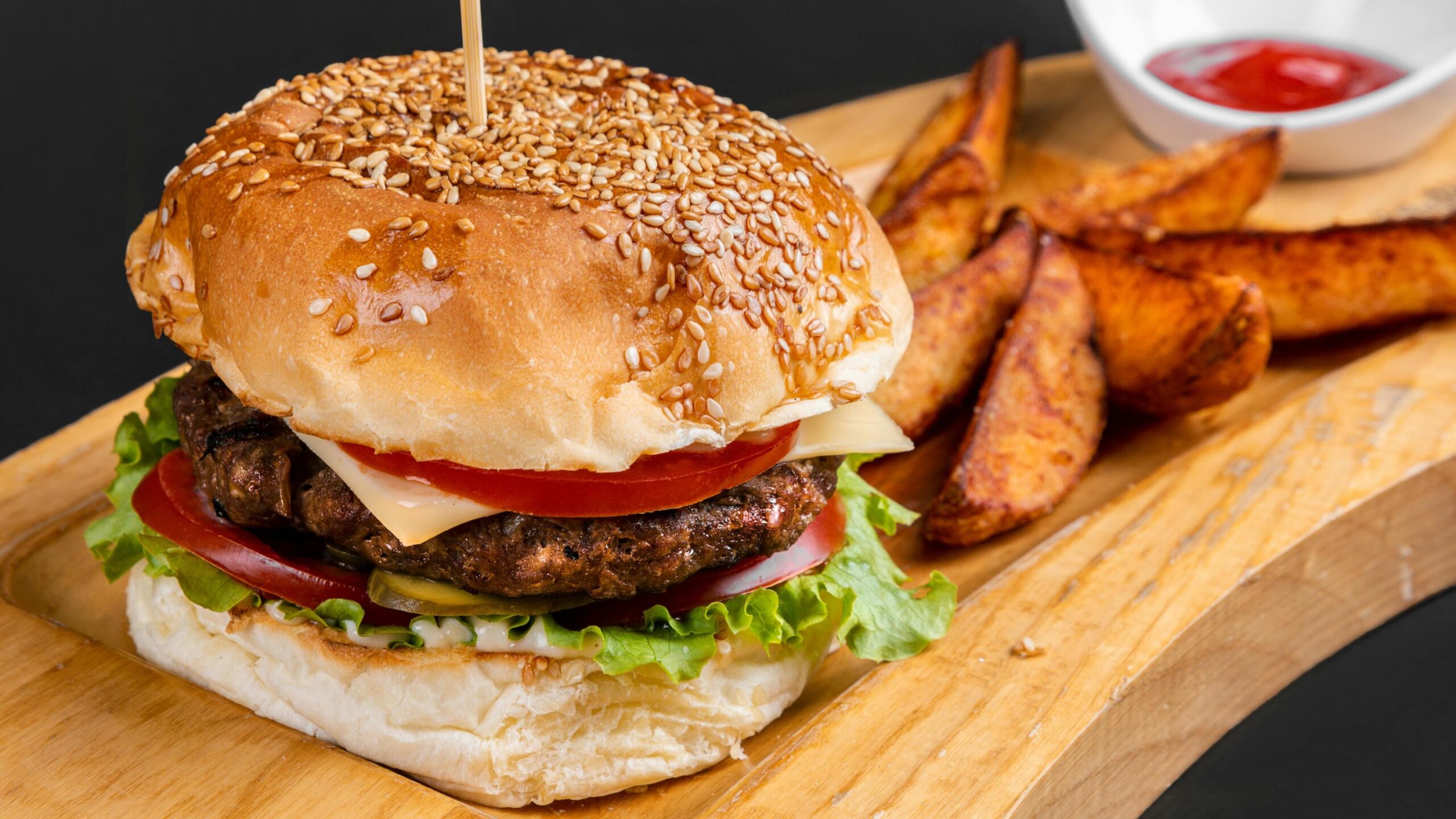 A hearty hamburger with a sesame seed bun is presented on a wooden board, stacked with layers of fresh lettuce, tomato, cheese, pickles, and a grilled beef patty. Golden-brown seasoned potato wedges and a small bowl of ketchup accompany the burger, all set against a simple dark background.