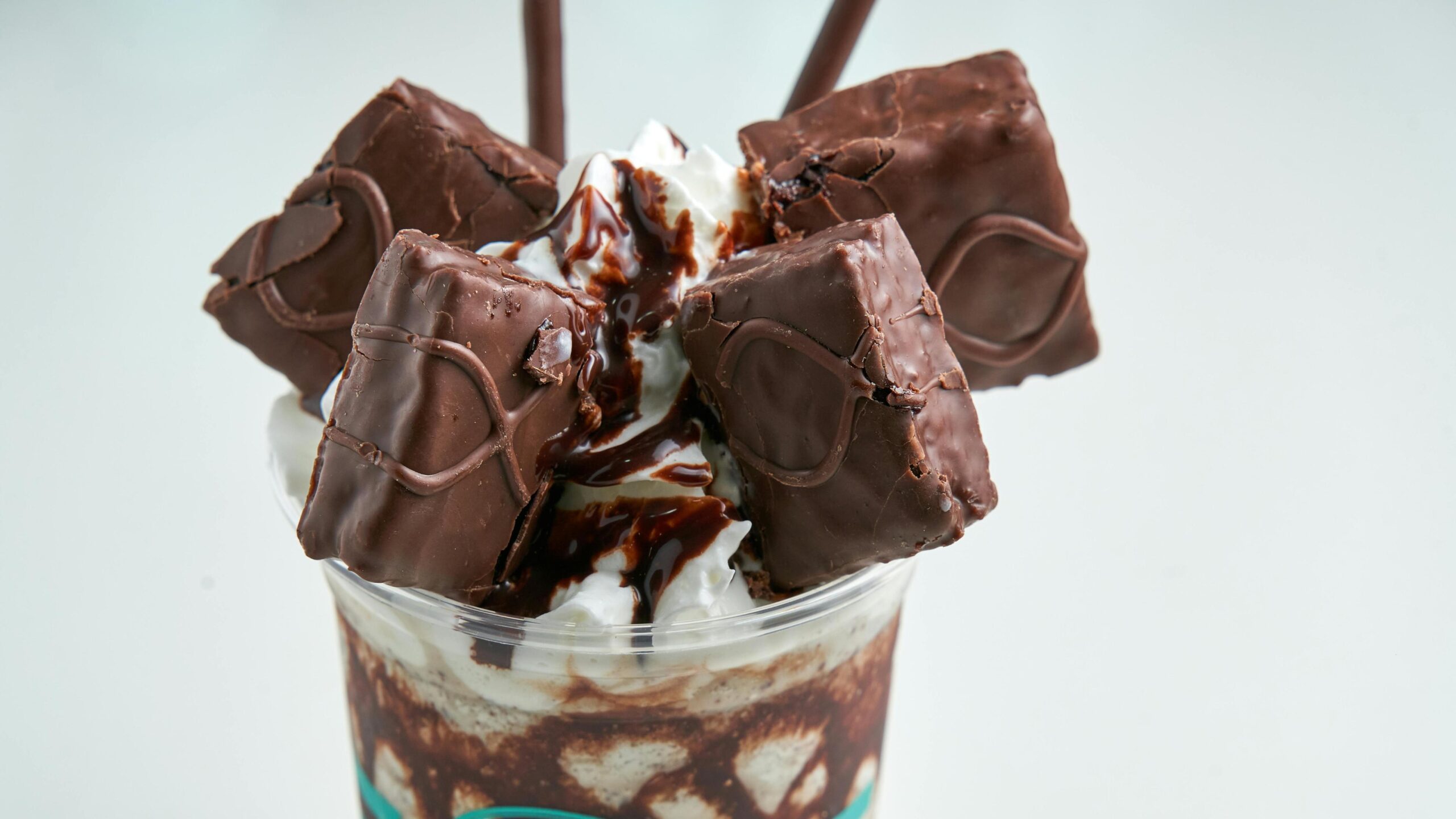 A decadent milkshake is piled high with whipped cream, rich chocolate drizzle, and several square chocolate-covered treats. Two thin chocolate sticks protrude from the top, adding a finishing touch to this indulgent dessert served in a clear cup with visible chocolate swirls.