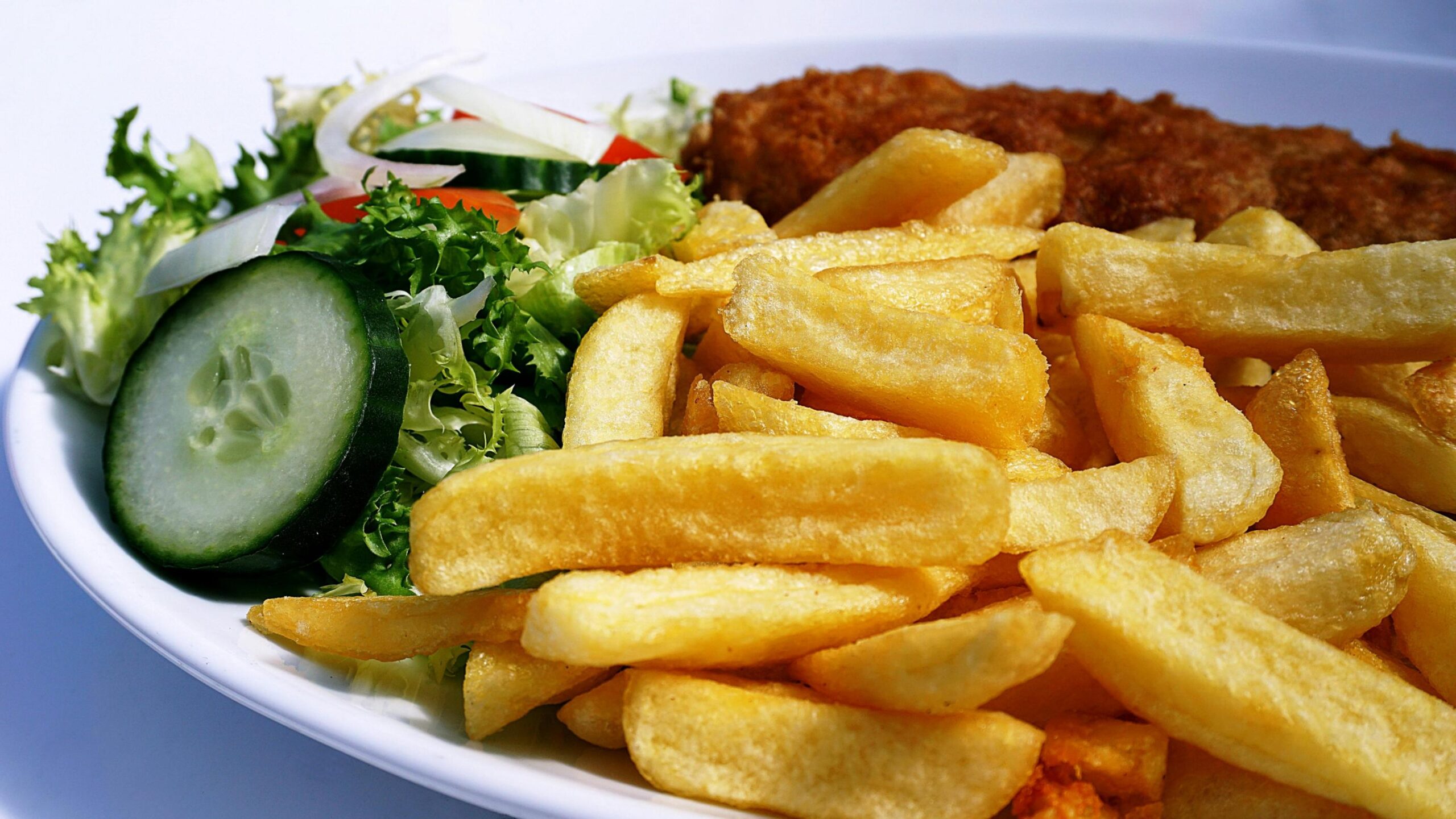 A close-up view shows a plate filled with golden, thick-cut french fries and a piece of crispy, breaded fish or meat in the background. A fresh side salad containing crisp lettuce, cucumber slices, and red onion sits to the left, adding a splash of color to the meal.