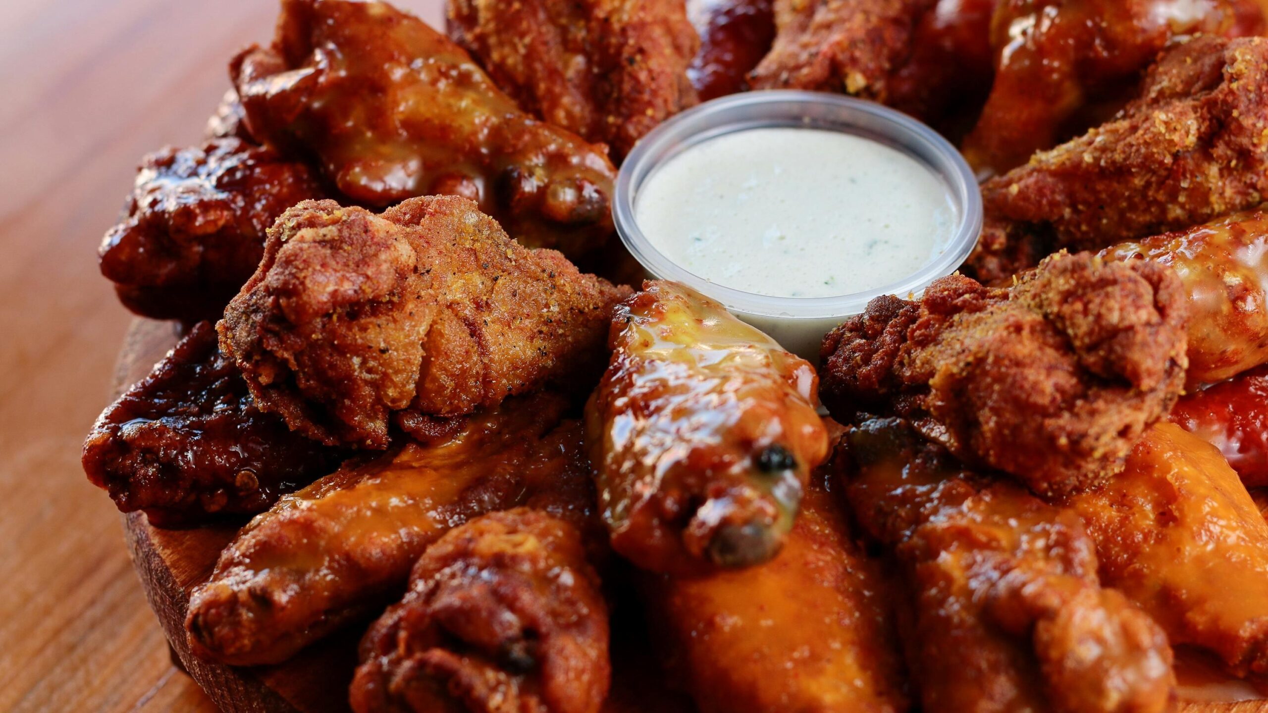 A wooden board presents a mouth-watering circle of chicken wings, showcasing a variety of textures from crispy and seasoned to glossy and sauced. Nestled in the center of the arrangement is a small plastic cup filled with a creamy white dipping sauce.