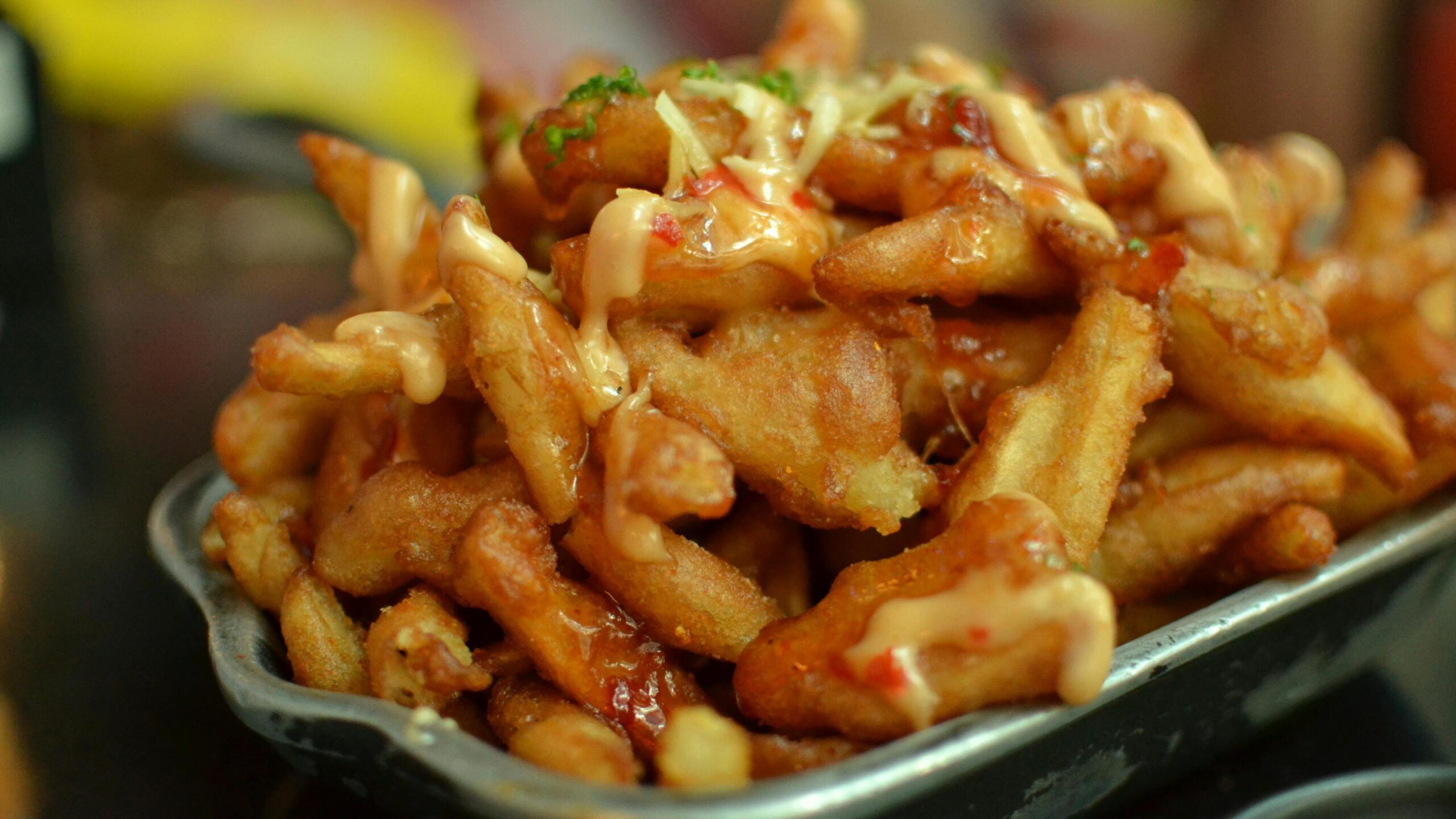 A metal tray is piled high with crispy, golden fried potato wedges drizzled with a creamy sauce and a glossy red glaze. Garnished with shredded toppings and fresh herbs, this indulgent appetizer is captured in a close-up shot that highlights its savory texture.
