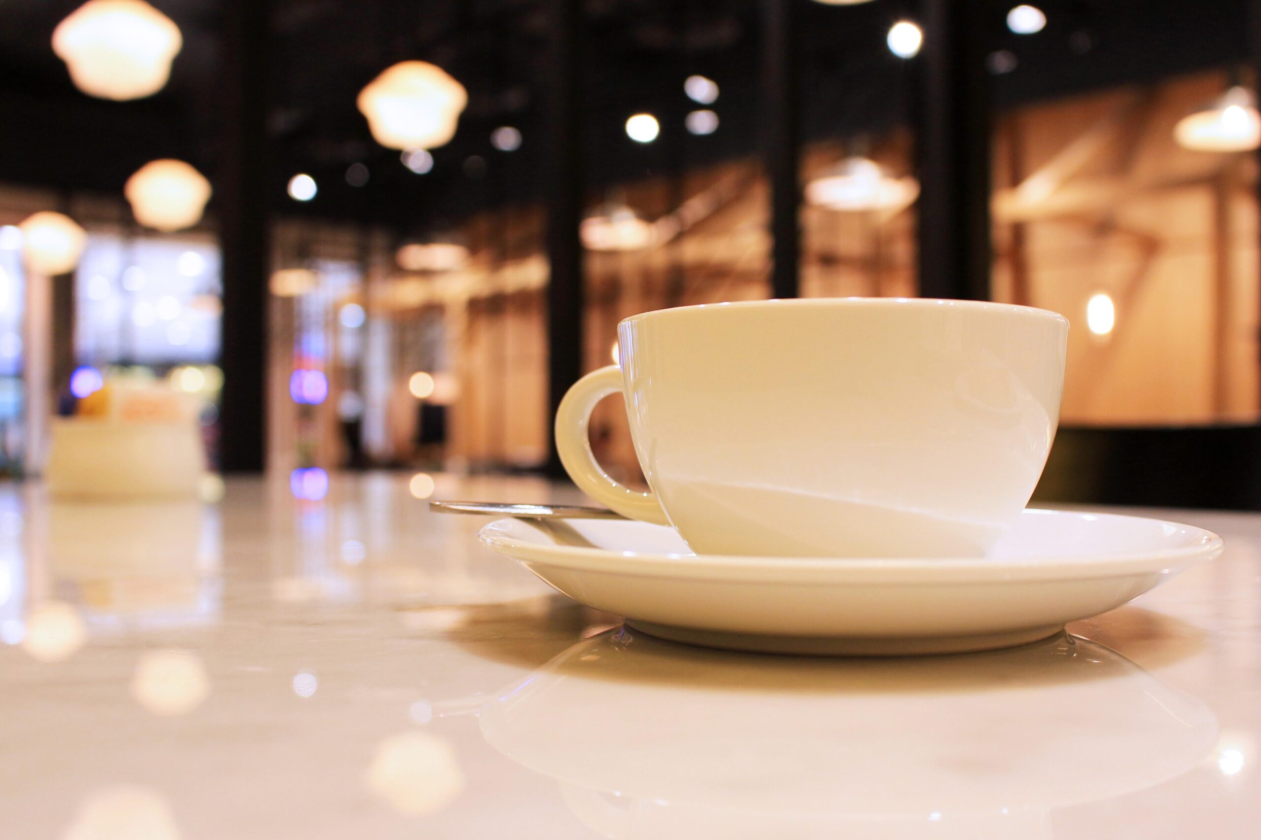 Tampines Coffee - 1 A white ceramic coffee cup and saucer sit on a glossy, reflective table, creating a sharp focal point in the foreground. The background features a softly blurred coffee shop interior with warm, glowing hanging lights that evoke a cozy and inviting atmosphere.