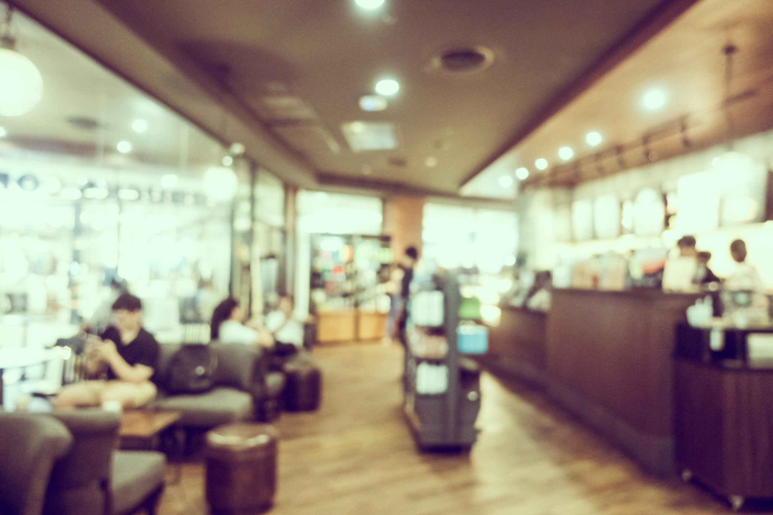 Tampines Coffee - 2 This abstract, defocused photograph depicts the interior of a bustling cafe or restaurant, characterized by a warm, sepia-toned color palette. Blurry figures of customers seated on the left and a service counter on the right are visible through the soft haze and bokeh lighting, creating a lively yet indistinct atmosphere.