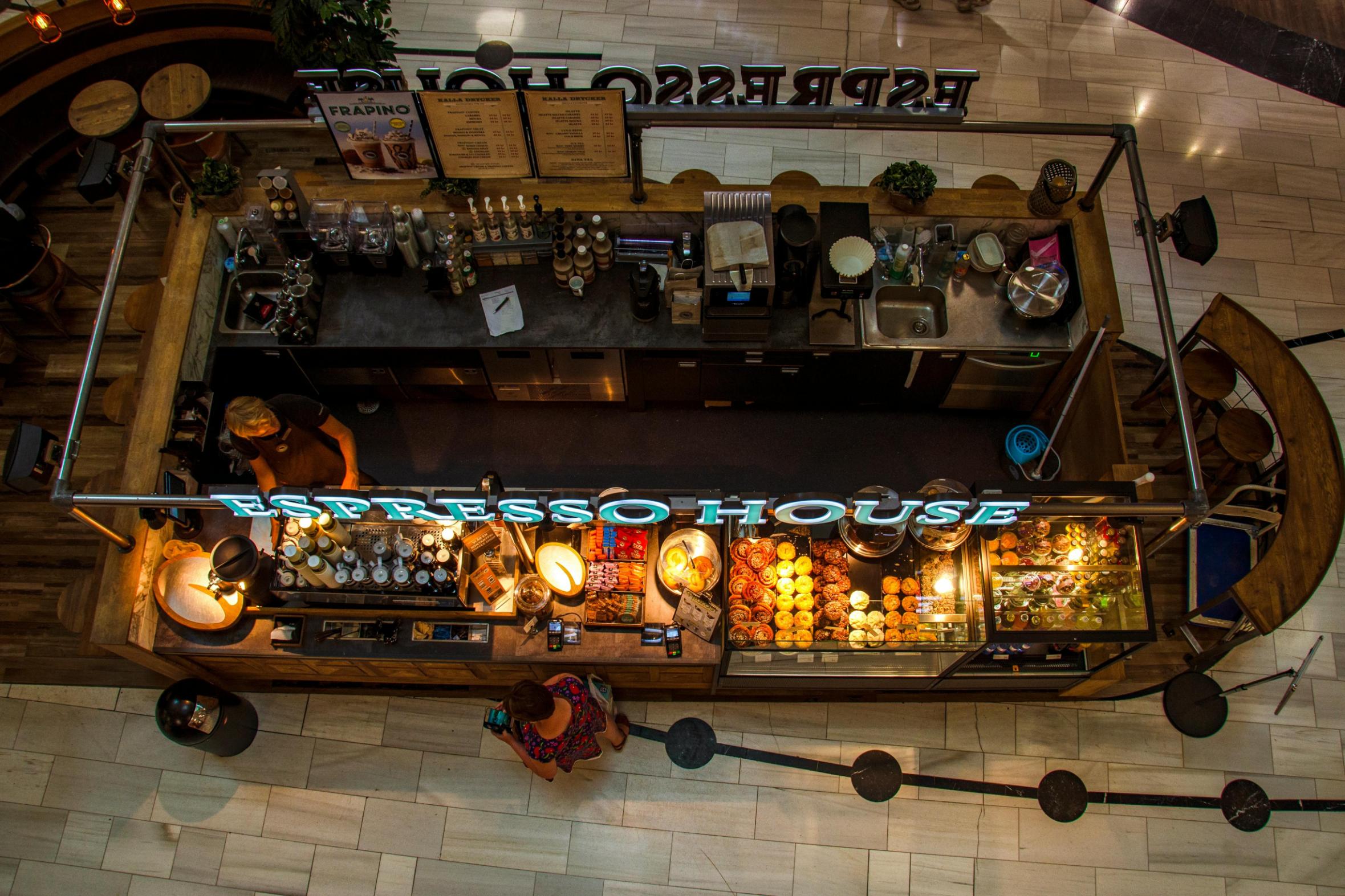 Tampines Coffee - 3 Captured from a bird's-eye view, this image shows a freestanding "Espresso House" kiosk situated on a tiled floor, featuring curved wooden counters and overhead menu boards. Inside the station, a barista works near a glass display case filled with pastries, while a customer stands at the counter beneath the bright teal signage.