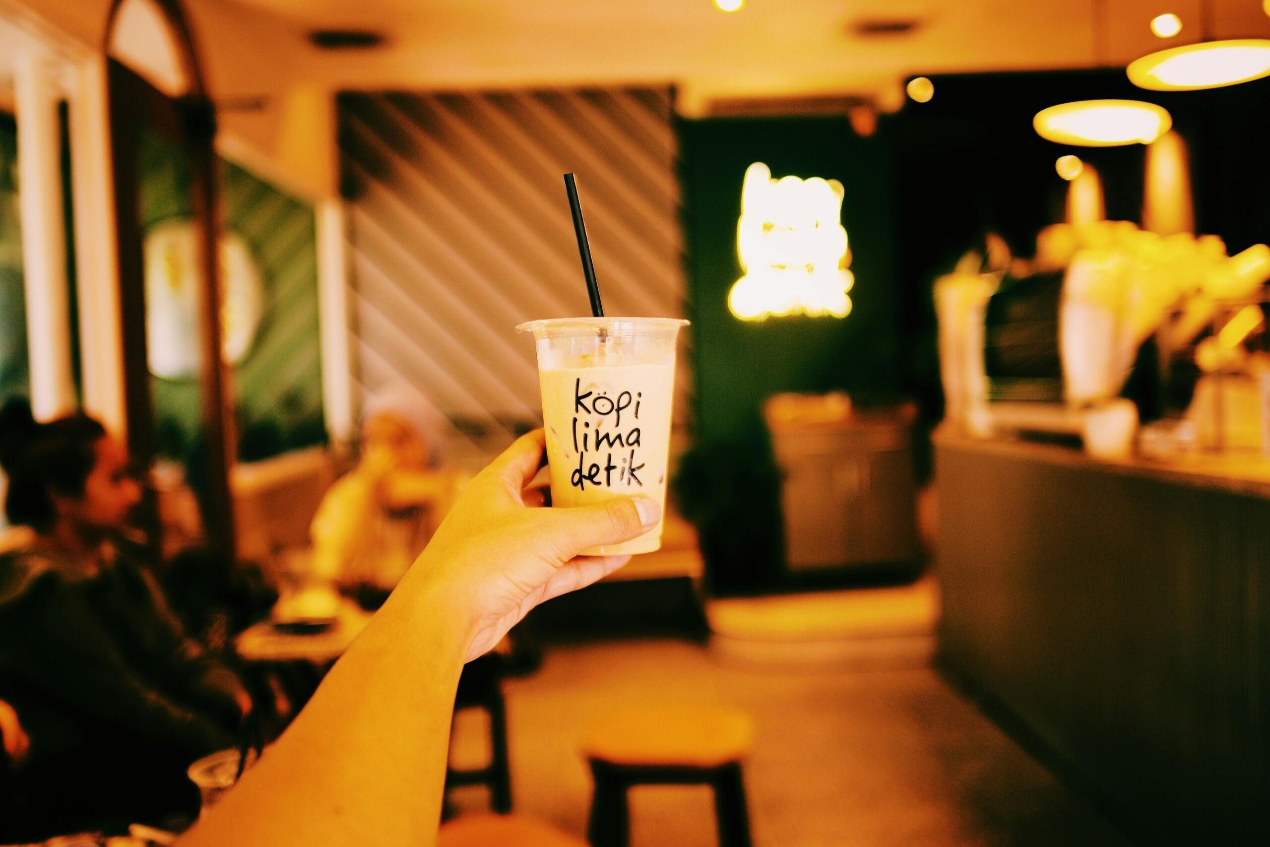 Tampines Coffee - 4 In the foreground, a hand holds up a clear plastic cup filled with an iced coffee beverage and a black straw, featuring the text "kopi lima detik" written on the side. The background reveals a warm, amber-lit cafe interior with blurred patrons seated on the left and a glowing neon sign on the back wall.