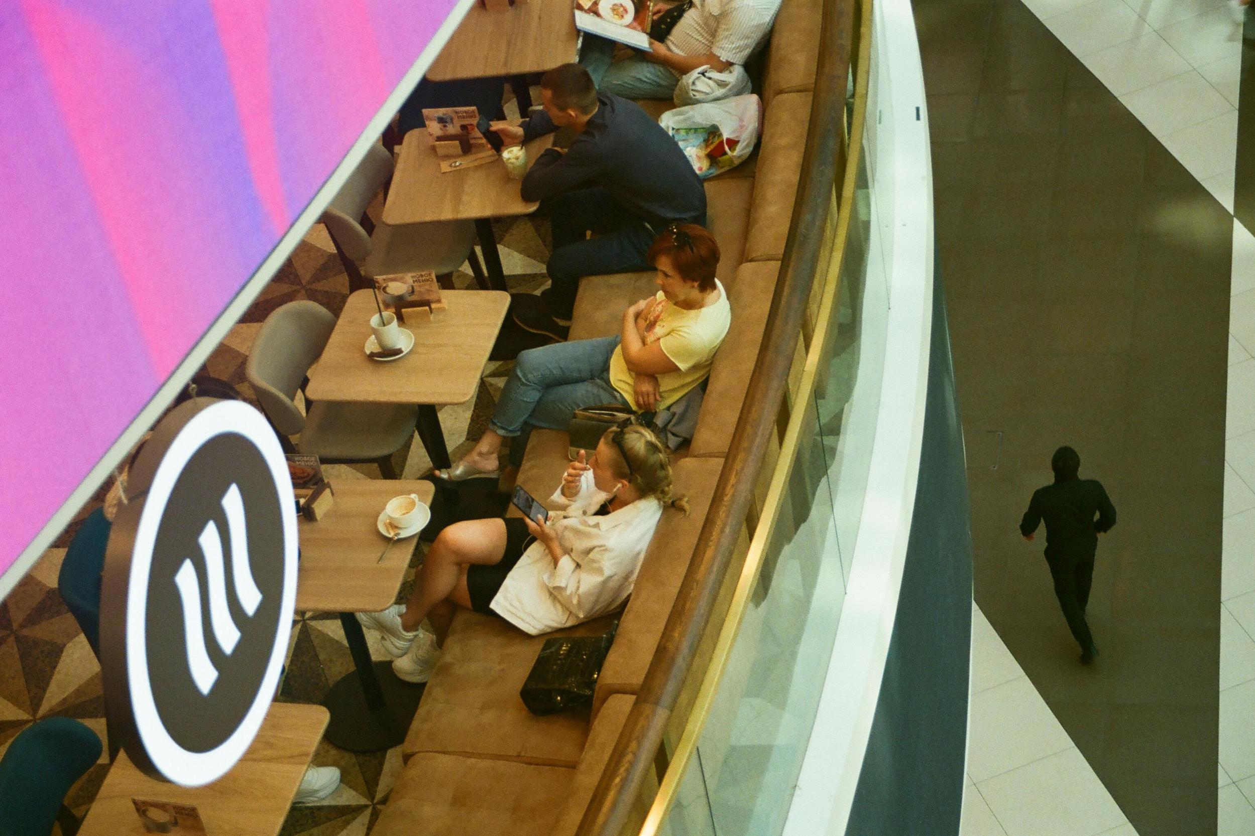 Tampines Coffee - 5 Taken from a high-angle perspective, this image shows a curved café seating area along a glass railing where several patrons relax at small wooden tables on a geometric patterned floor. In the foreground, a prominent circular sign with a wavy logo hangs near the diners, while a lone figure dressed in black walks across the polished, tiled corridor to the right.