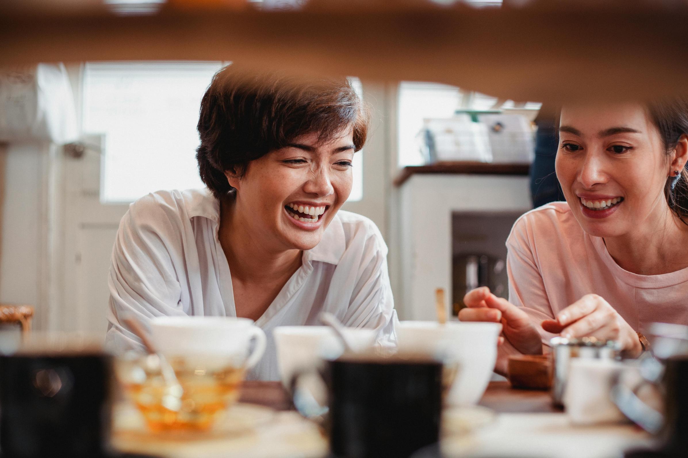 Tampines Coffee - 6 Two smiling women are captured in a candid moment of laughter while seated together at a table. In the foreground, out-of-focus cups and jars frame the scene, emphasizing the cozy and intimate atmosphere of their conversation.