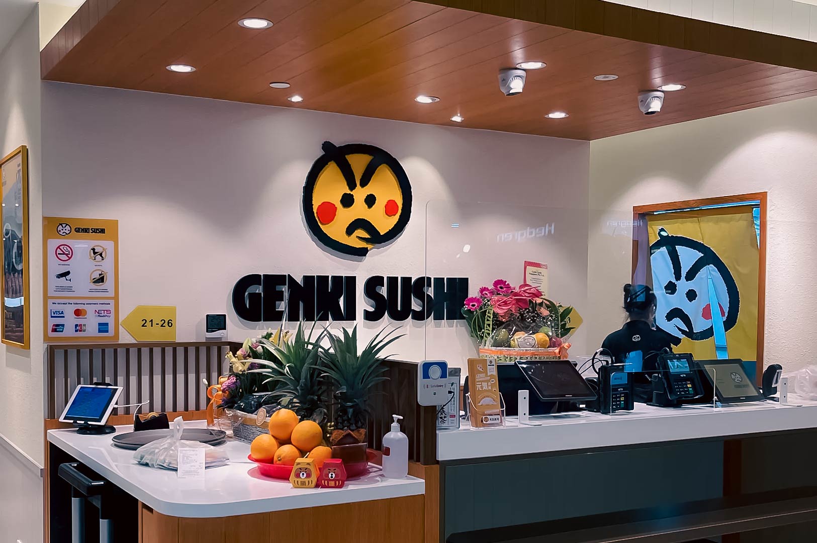 Genki Sushi Singapore counter with fresh fruit display, flowers, digital ordering screens, and the iconic yellow sushi logo.