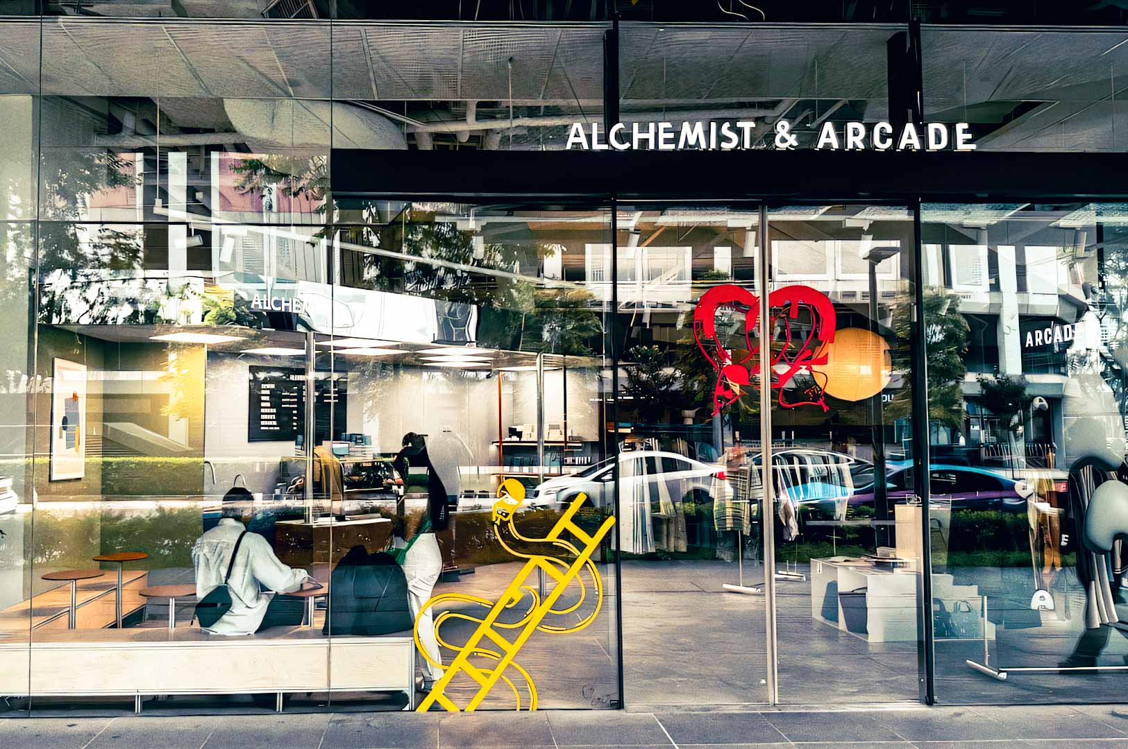Exterior of Alchemist & Arcade at Funan, showcasing its glass storefront, modern café interior, visible seating area, and decorative window graphics.